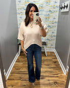 A woman takes a mirror selfie in a fitting room wearing a light beige short-sleeve knit top with a curved hem, dark blue flare jeans, and leopard print heels.