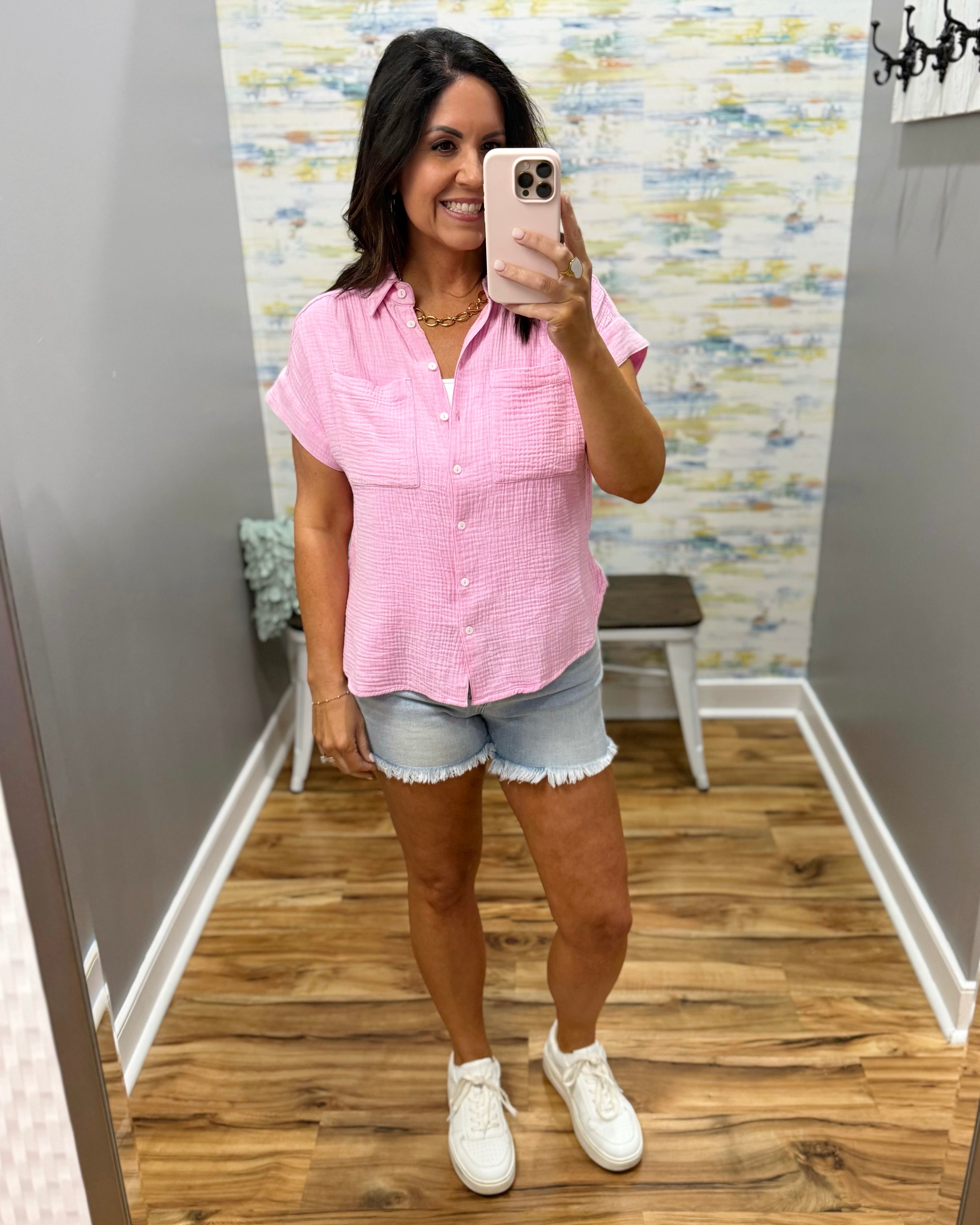Woman taking a mirror selfie wearing a pink checkered shirt and denim shorts.
