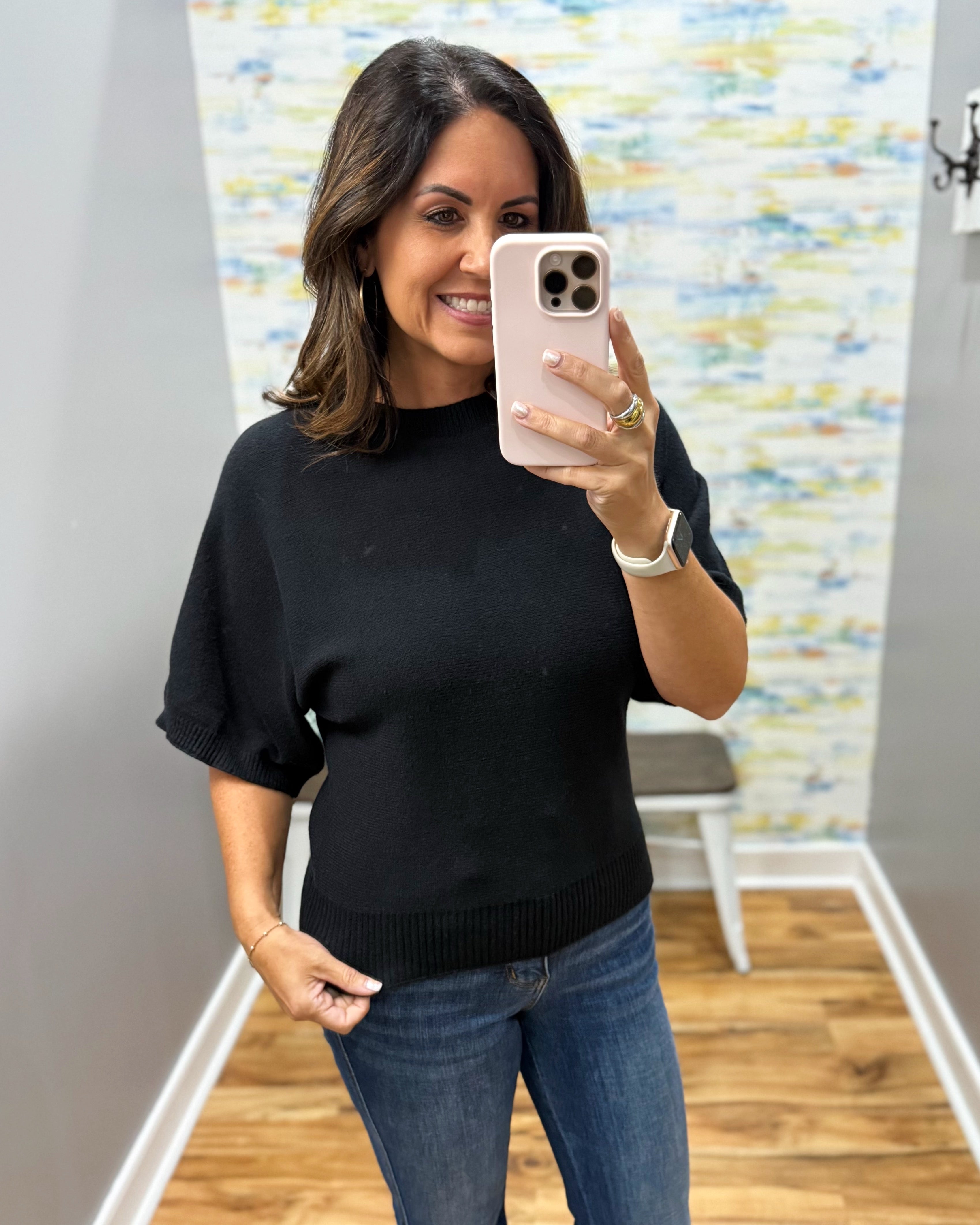 A woman takes a mirror selfie wearing a black short-sleeve knit sweater and blue jeans in a fitting room with wood floors and a patterned wall.