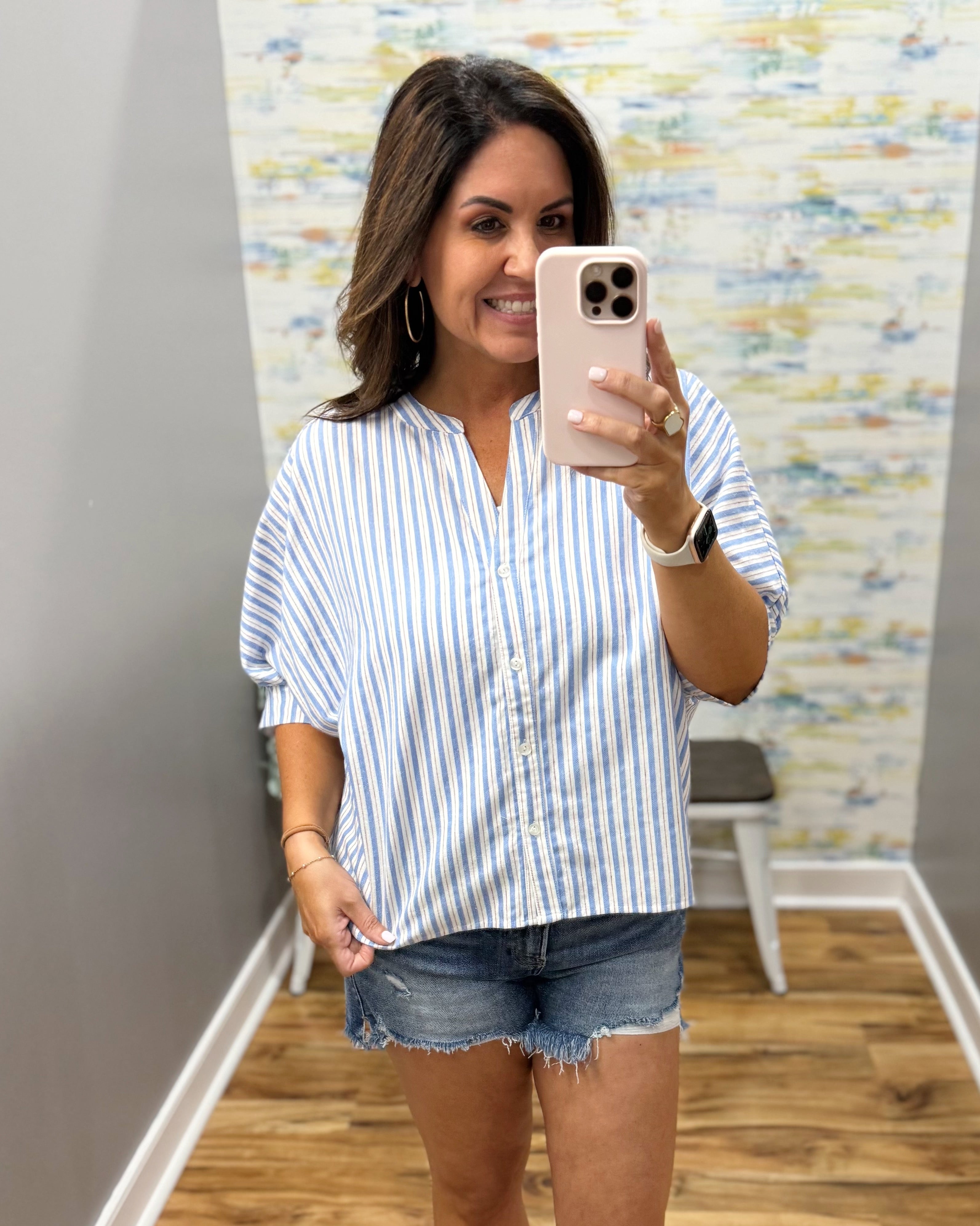 Woman taking a mirror selfie wearing a striped shirt and denim shorts in a room with a colorful wall.