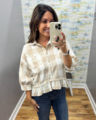 A woman taking a mirror selfie wearing a beige and white checkered ruffled blouse with dark blue jeans.