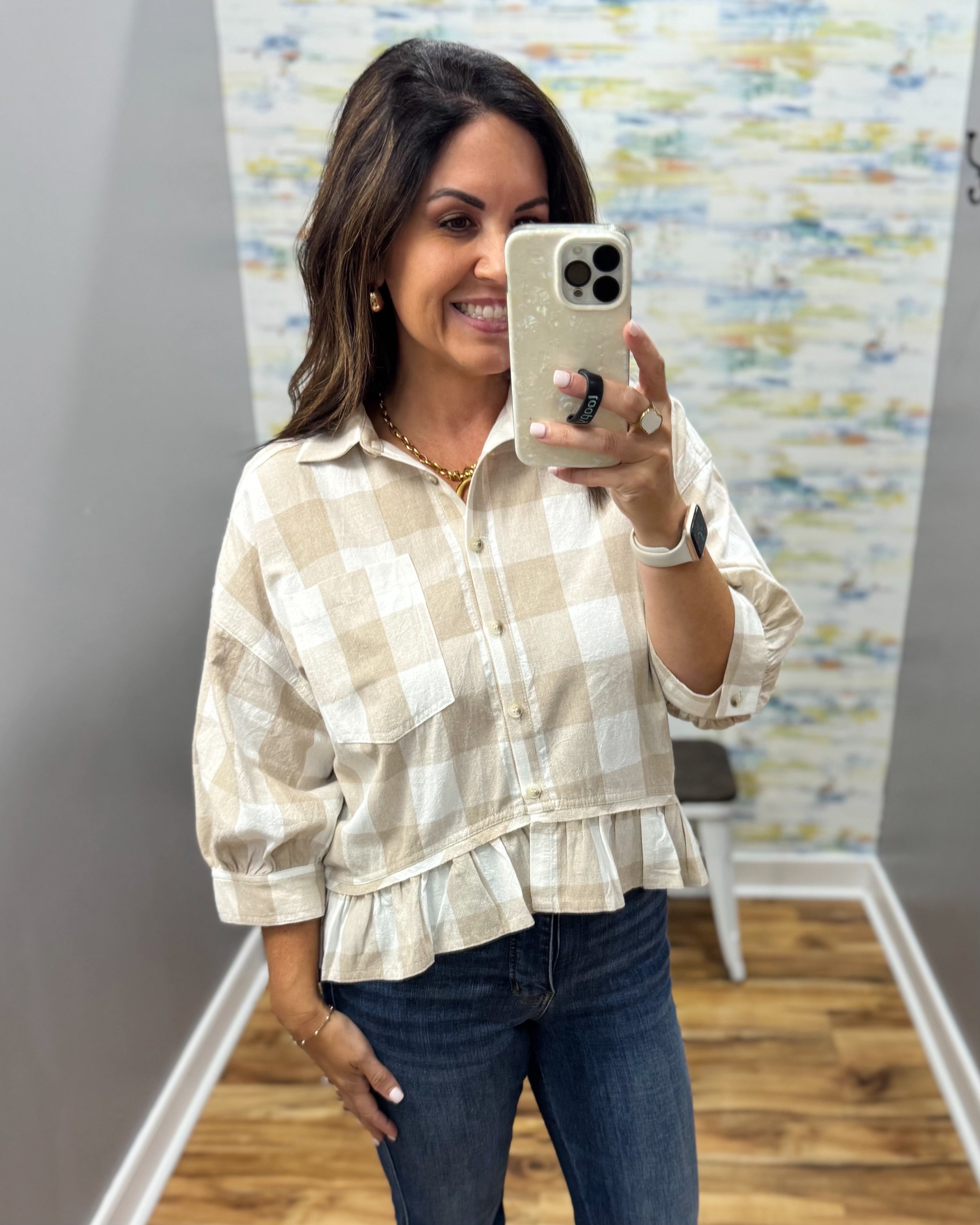 A woman taking a mirror selfie wearing a beige and white checkered ruffled blouse with dark blue jeans.