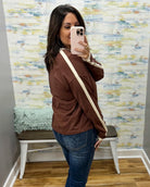 A woman takes a mirror selfie wearing a brown long-sleeve athletic jacket with cream stripes on the sleeves and a matching brown crossbody bag, paired with blue jeans.