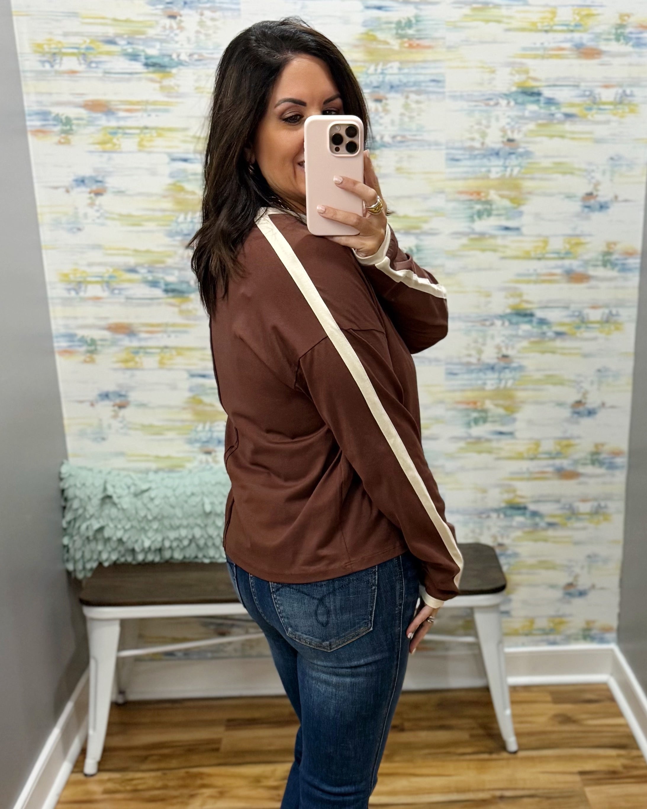 A woman takes a mirror selfie wearing a brown long-sleeve athletic jacket with cream stripes on the sleeves and a matching brown crossbody bag, paired with blue jeans.