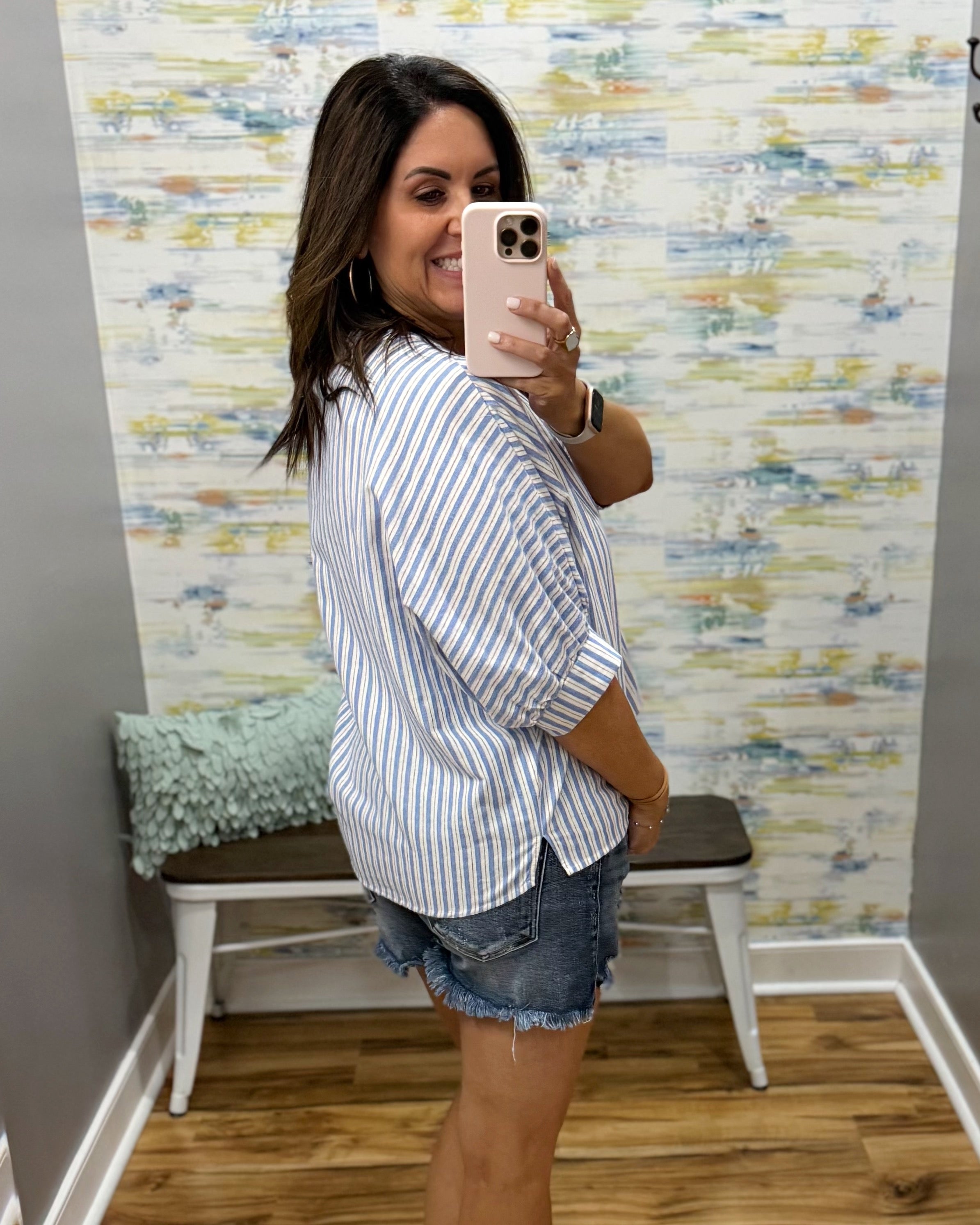 Woman taking a mirror selfie wearing a striped shirt and denim shorts in a room with colorful wall paper.