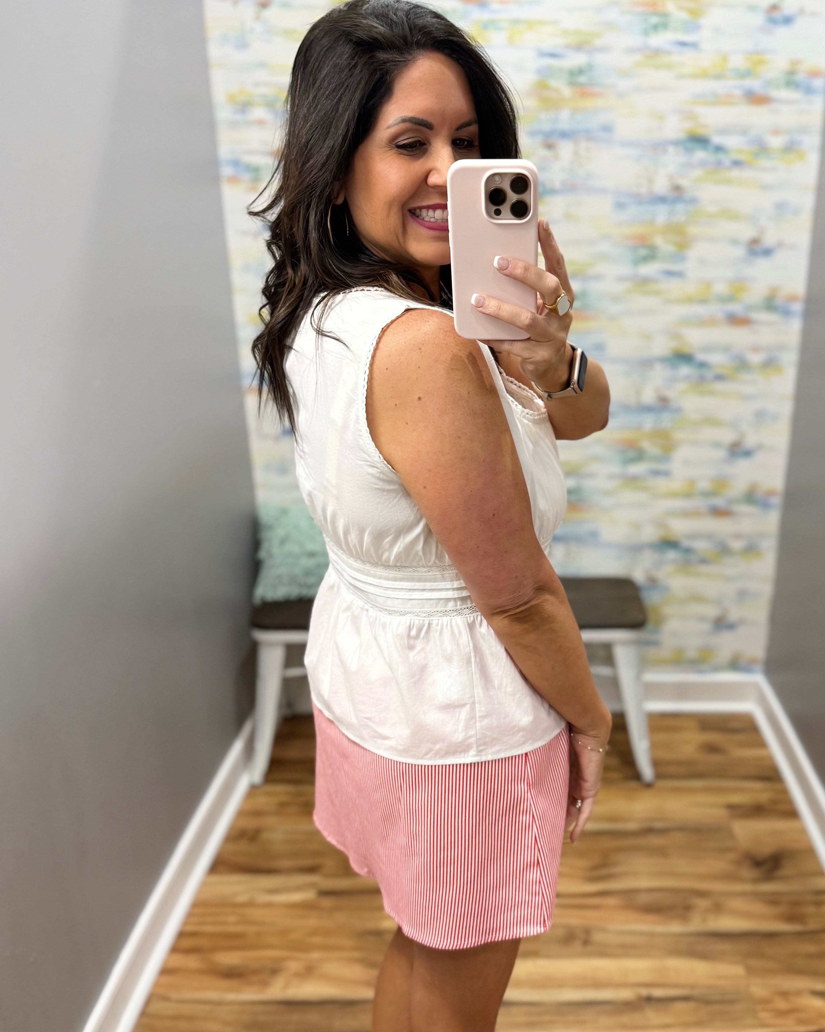 Woman taking a mirror selfie wearing a white sleeveless top and pink skirt.