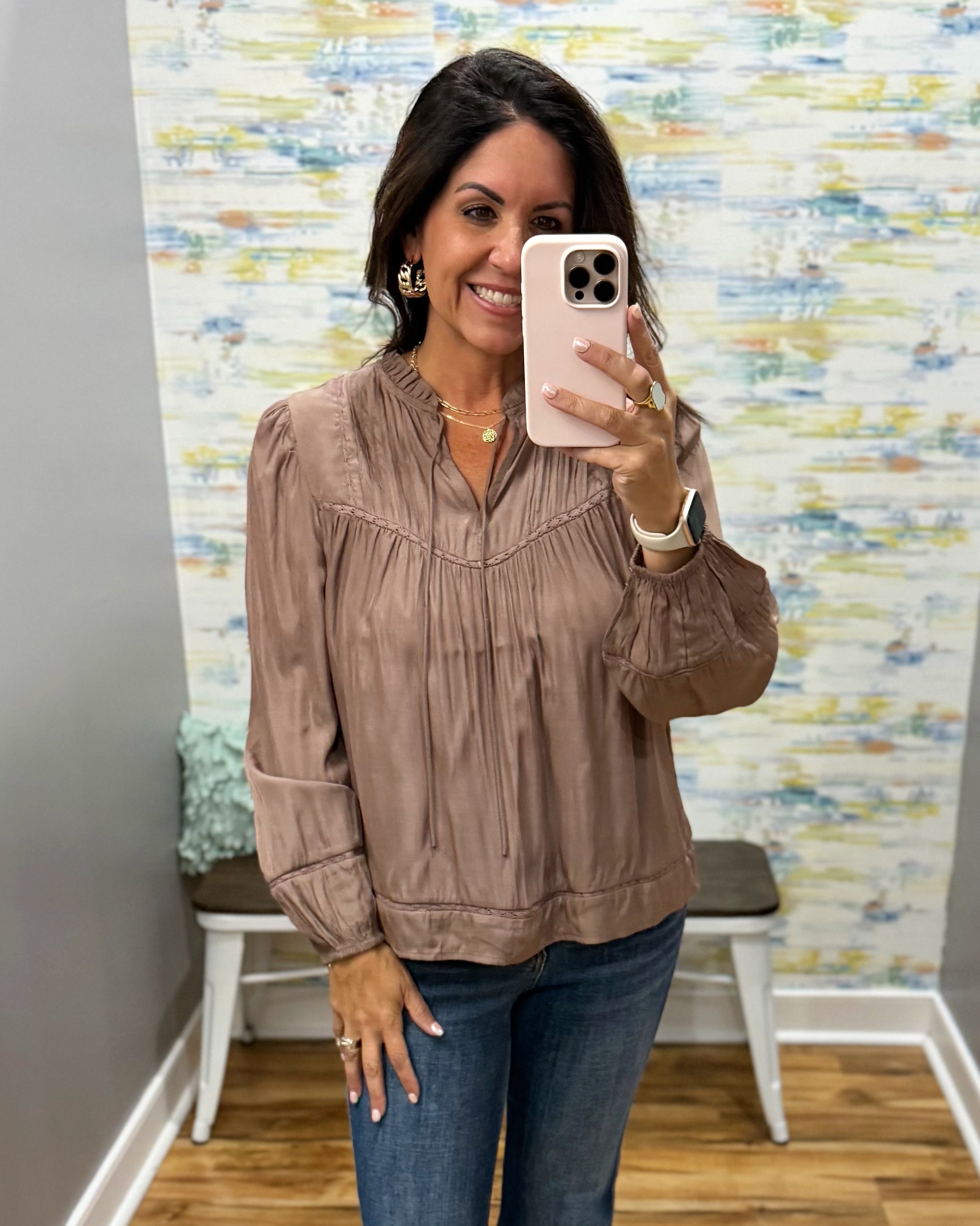 A woman takes a mirror selfie wearing a taupe brown long-sleeve blouse with a smocked chest and gathered cuffs, paired with blue jeans, in front of a patterned wallpaper background.