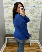 A woman takes a mirror selfie wearing a vibrant royal blue chunky knit sweater and light blue jeans in front of a patterned wallpapered wall.