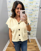 A woman takes a mirror selfie wearing a cream-colored short-sleeve knit cardigan with dark buttons and blue jeans in a fitting room.