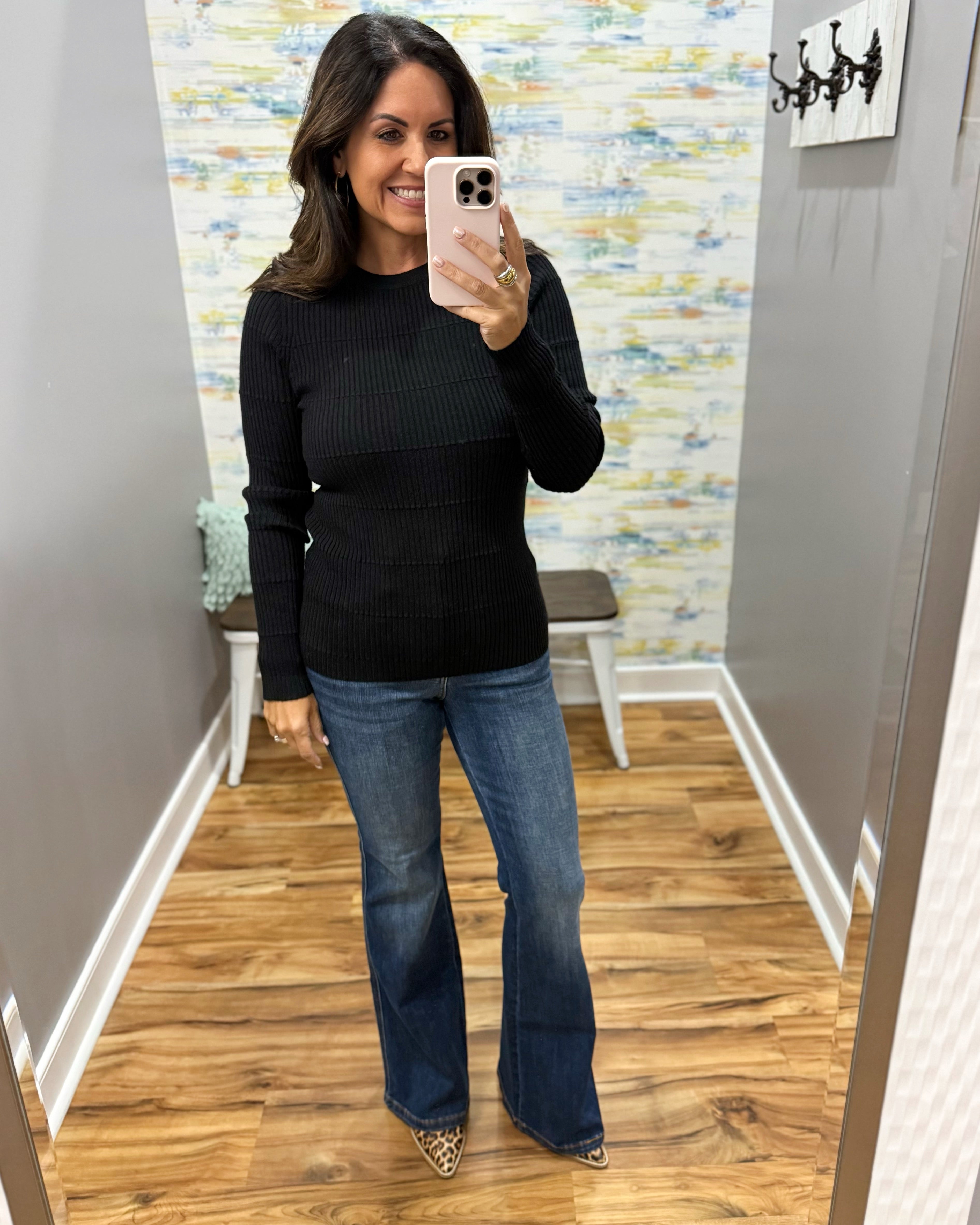 A woman takes a mirror selfie showing a black ribbed long-sleeve top and blue flare jeans in a fitting room with wood floors and a patterned wallpaper background.