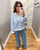 A woman takes a mirror selfie in a boutique showing a blue and white striped quarter-zip knit sweater and wide-leg blue jeans.