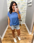Woman taking a mirror selfie wearing a blue top and denim shorts in a room with a textured wall.