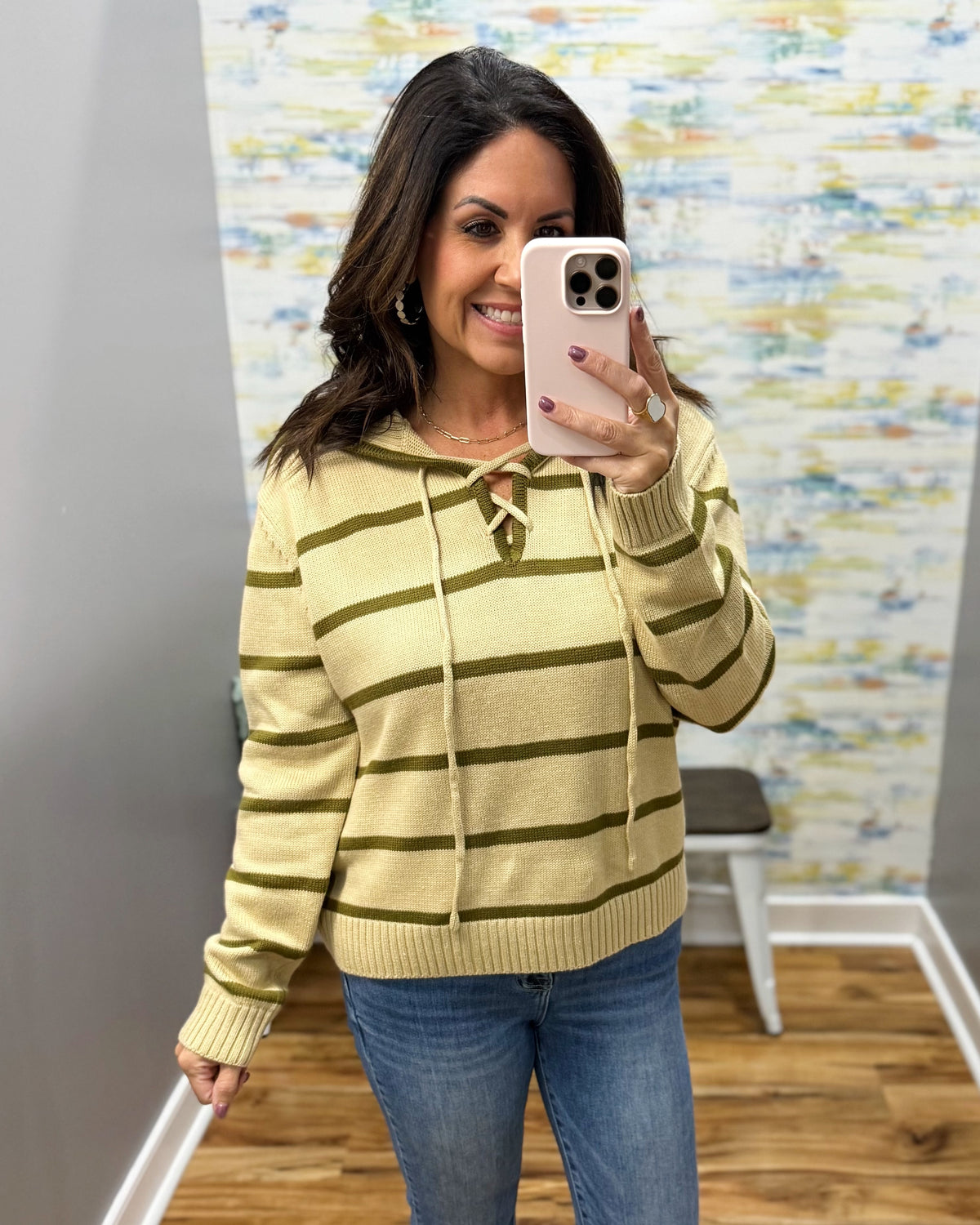 A woman takes a mirror selfie wearing a taupe and olive green striped knit hoodie with drawstrings and a front keyhole cutout, paired with blue jeans.