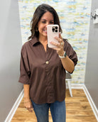 A woman takes a mirror selfie wearing a brown button-down shirt with rolled sleeves and blue jeans in a fitting room with wood floors and a patterned wall.