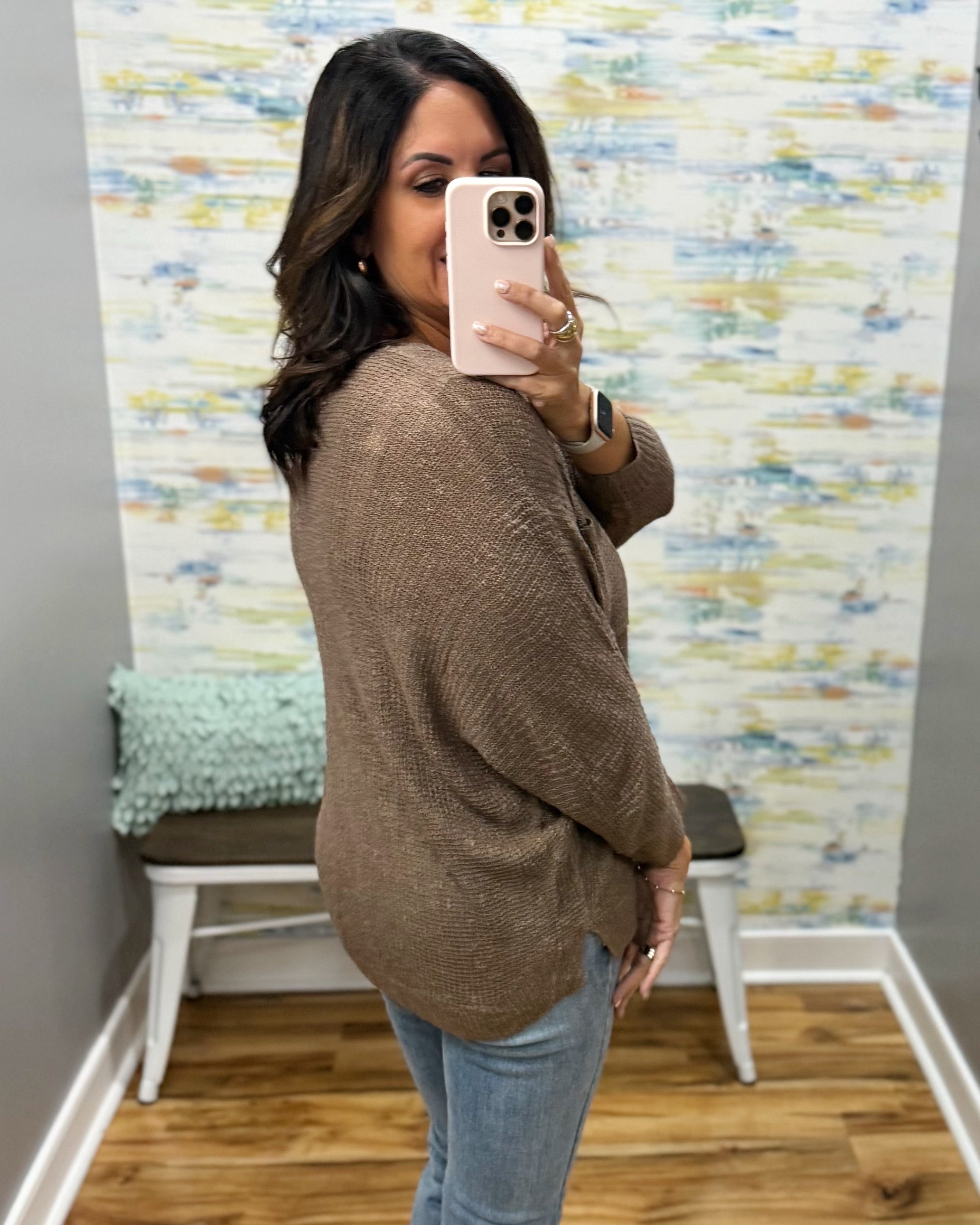 A woman takes a mirror selfie wearing a brown textured knit sweater and light blue jeans in a room with a patterned wallpaper background.