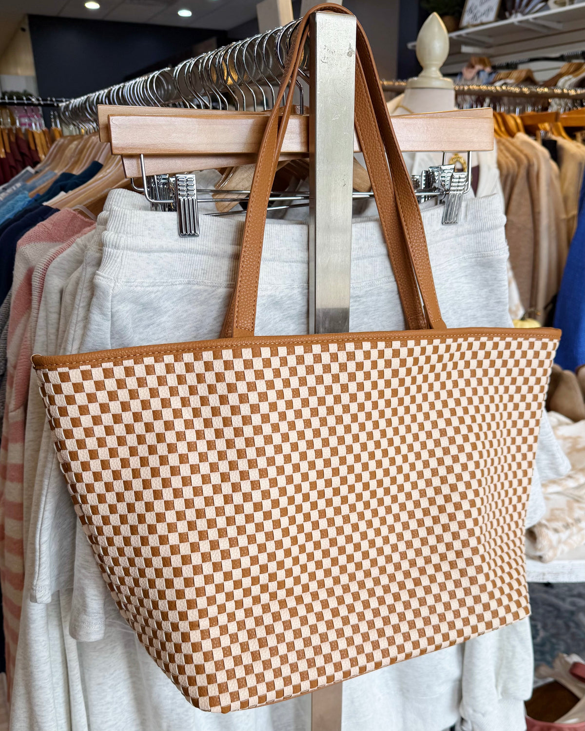A large woven tote bag with a tan and cream checkerboard pattern and brown shoulder straps, hanging on a clothing rack in a retail store.