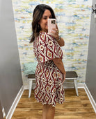 A woman takes a mirror selfie in a fitting room wearing a short-sleeve, knee-length dress with a red, beige, and white geometric diamond pattern.