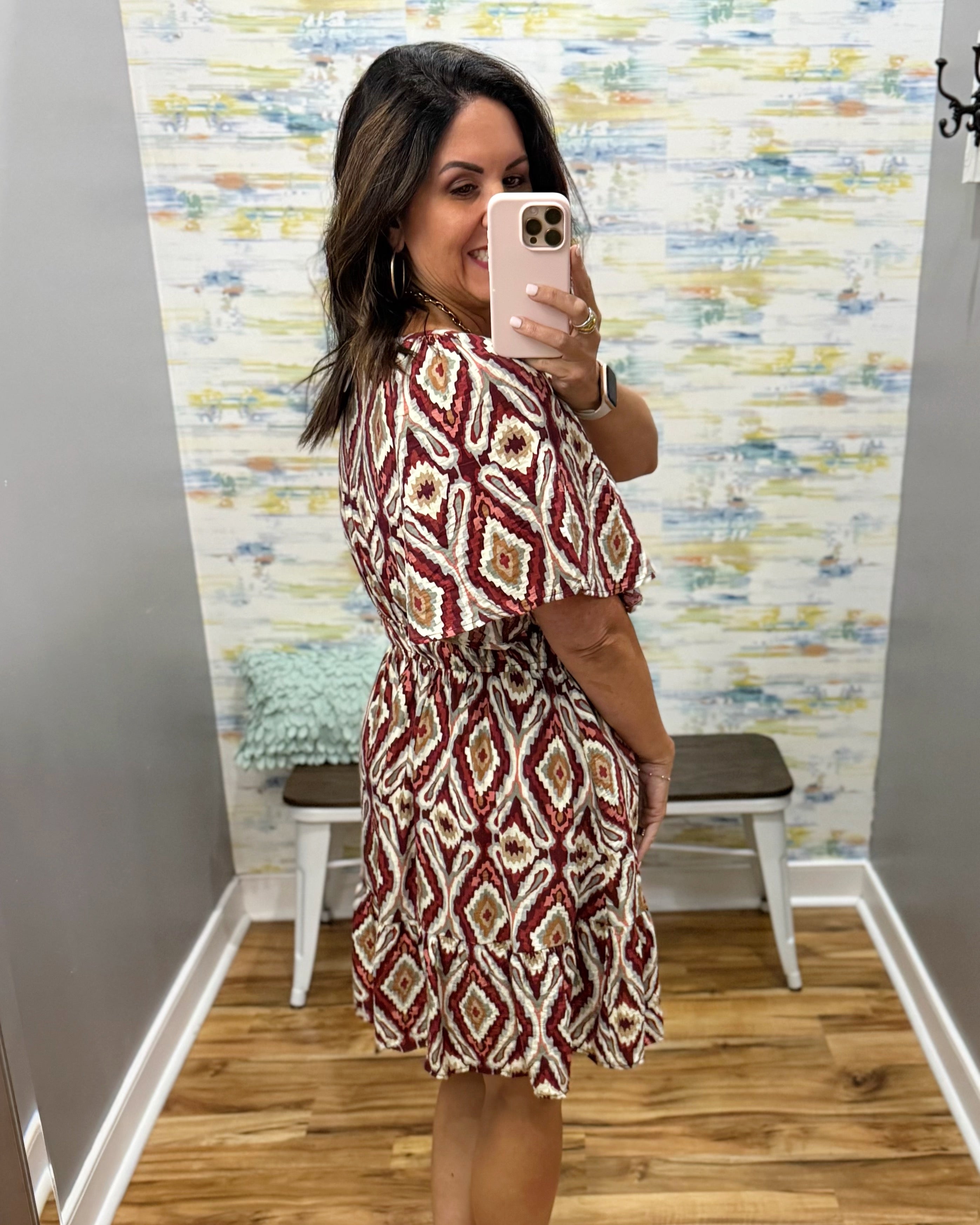 A woman takes a mirror selfie in a fitting room wearing a short-sleeve, knee-length dress with a red, beige, and white geometric diamond pattern.