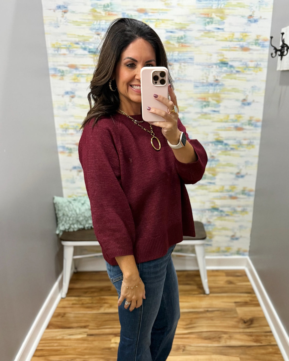 A woman takes a mirror selfie wearing a maroon knit sweater and blue jeans in a home setting with a patterned wallpaper background.