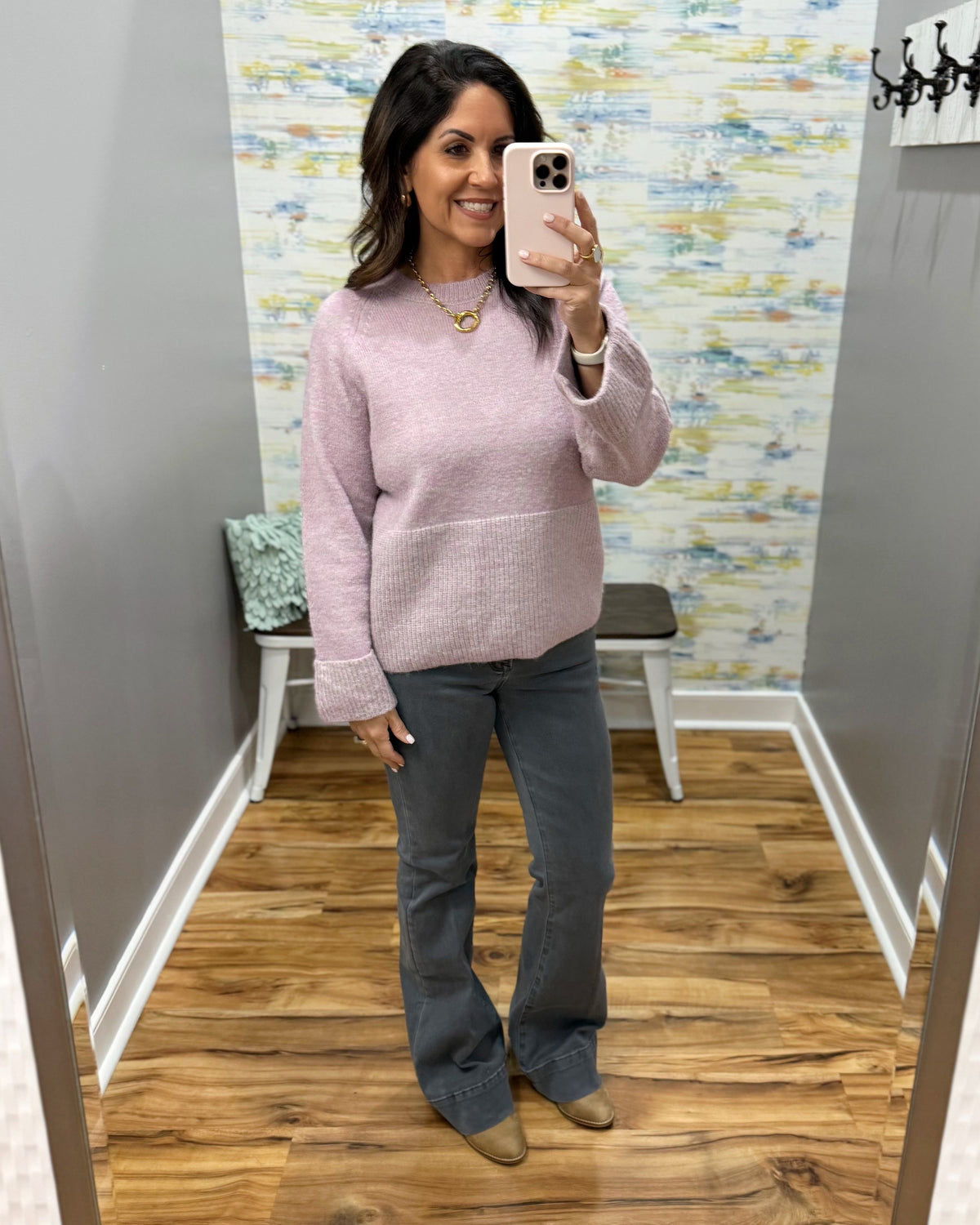 A woman takes a mirror selfie in a fitting room wearing a light pink knit sweater and dark gray flared pants.