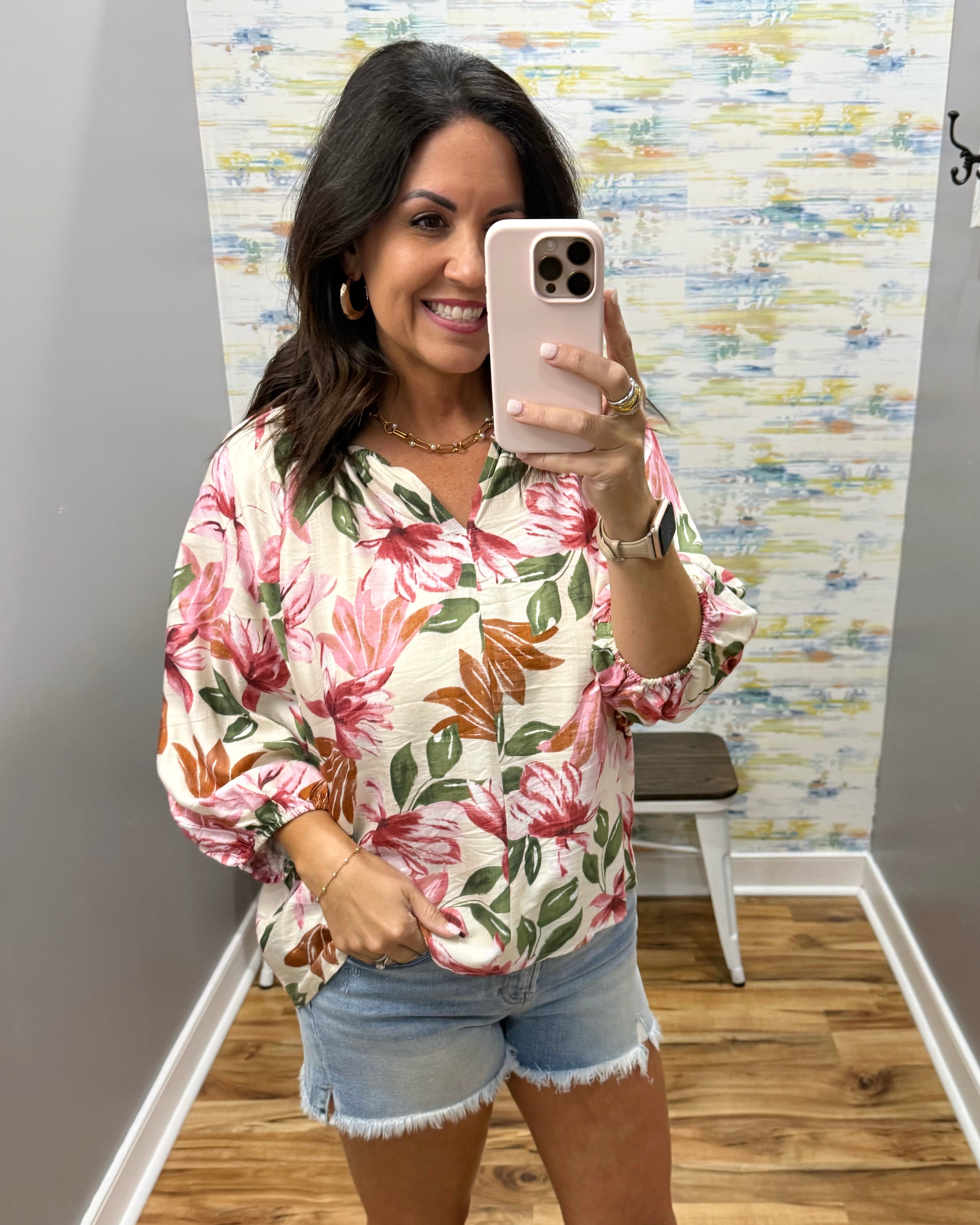 Woman taking a mirror selfie wearing a floral blouse and denim shorts in a room with colorful wall art.