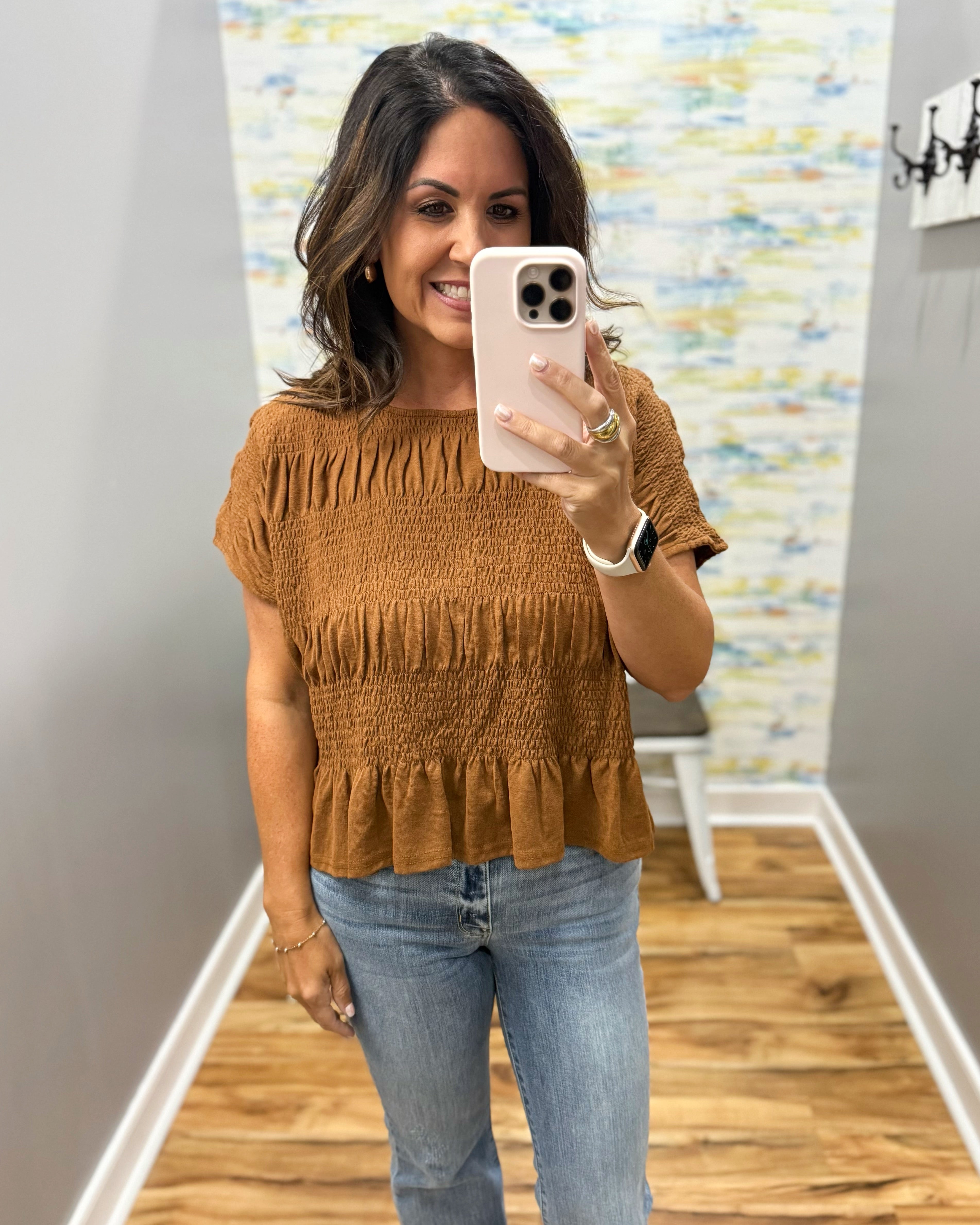 A woman taking a mirror selfie in a boutique, wearing a rust-brown smocked short-sleeve top and light-wash blue jeans.