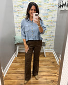 A woman takes a mirror selfie wearing a light blue denim ruffle-sleeve shirt and dark brown wide-leg pants in a home fitting room with wood floors and abstract wallpaper.