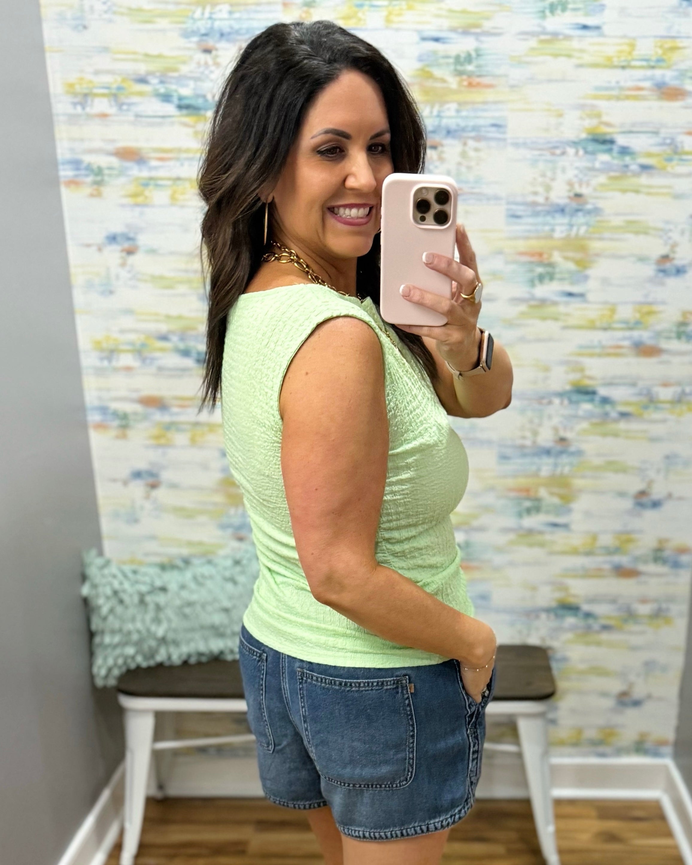 Woman wearing a light green top and denim shorts in a room with a textured wall.