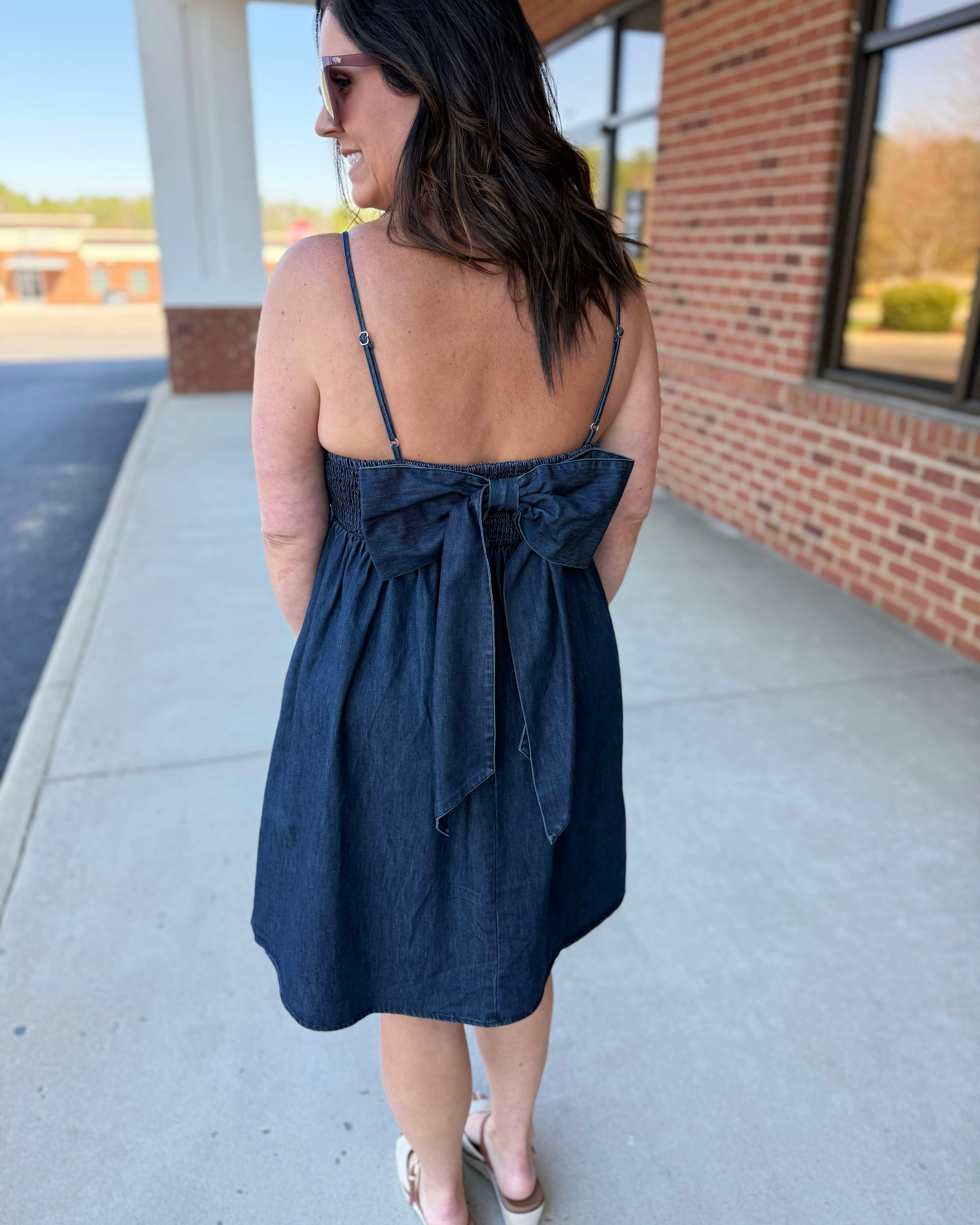 Woman wearing a black dress with a bow detail, standing on a sidewalk.