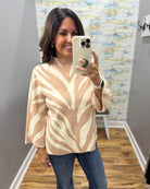 A woman takes a mirror selfie wearing a beige and cream knit sweater with a leaf-like pattern, paired with blue jeans, in an indoor hallway.