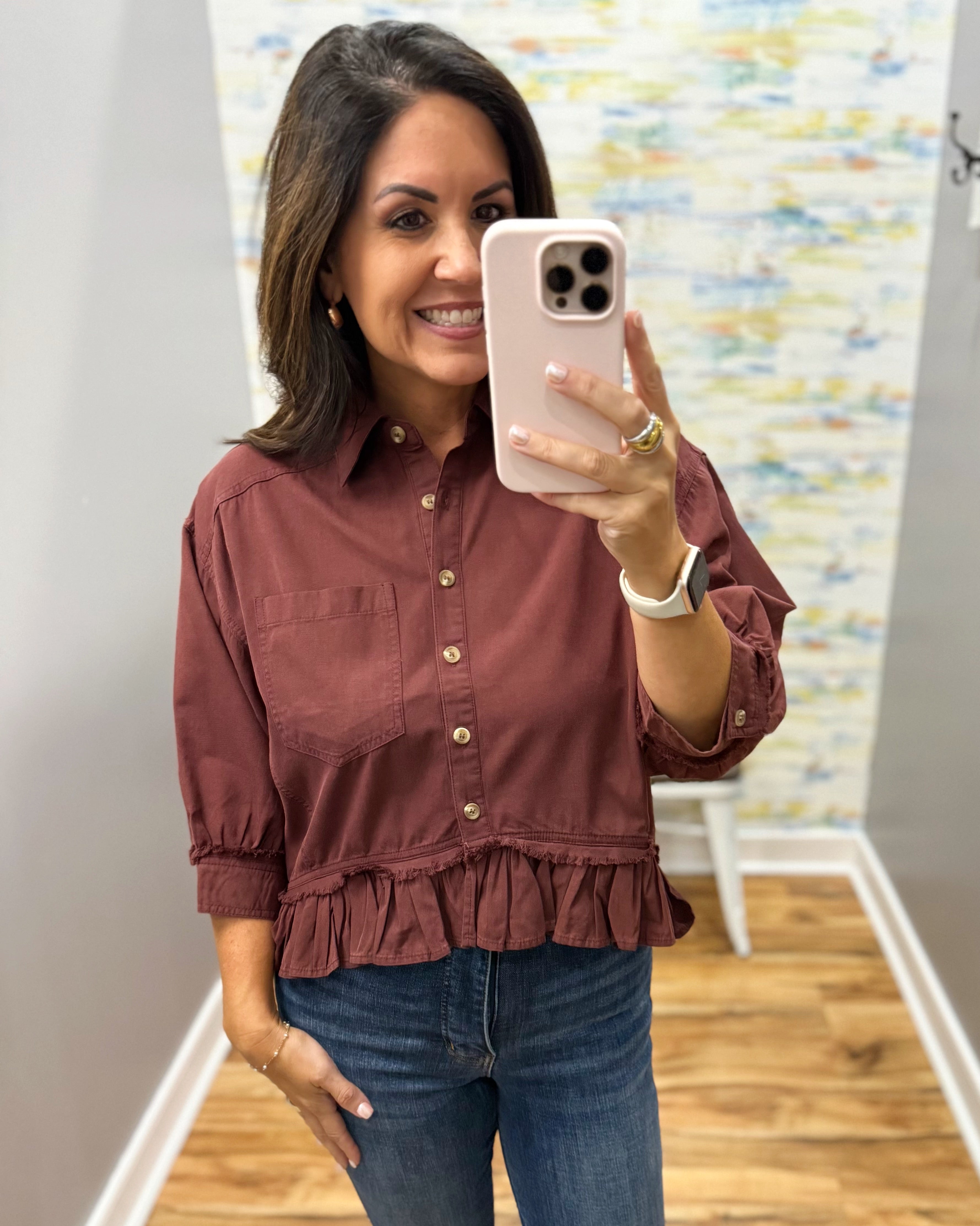 A woman takes a mirror selfie wearing a rust-colored, long-sleeve button-up blouse with a ruffled hem and blue jeans.