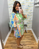 Woman taking a mirror selfie wearing a colorful floral dress in a room with a patterned wall.