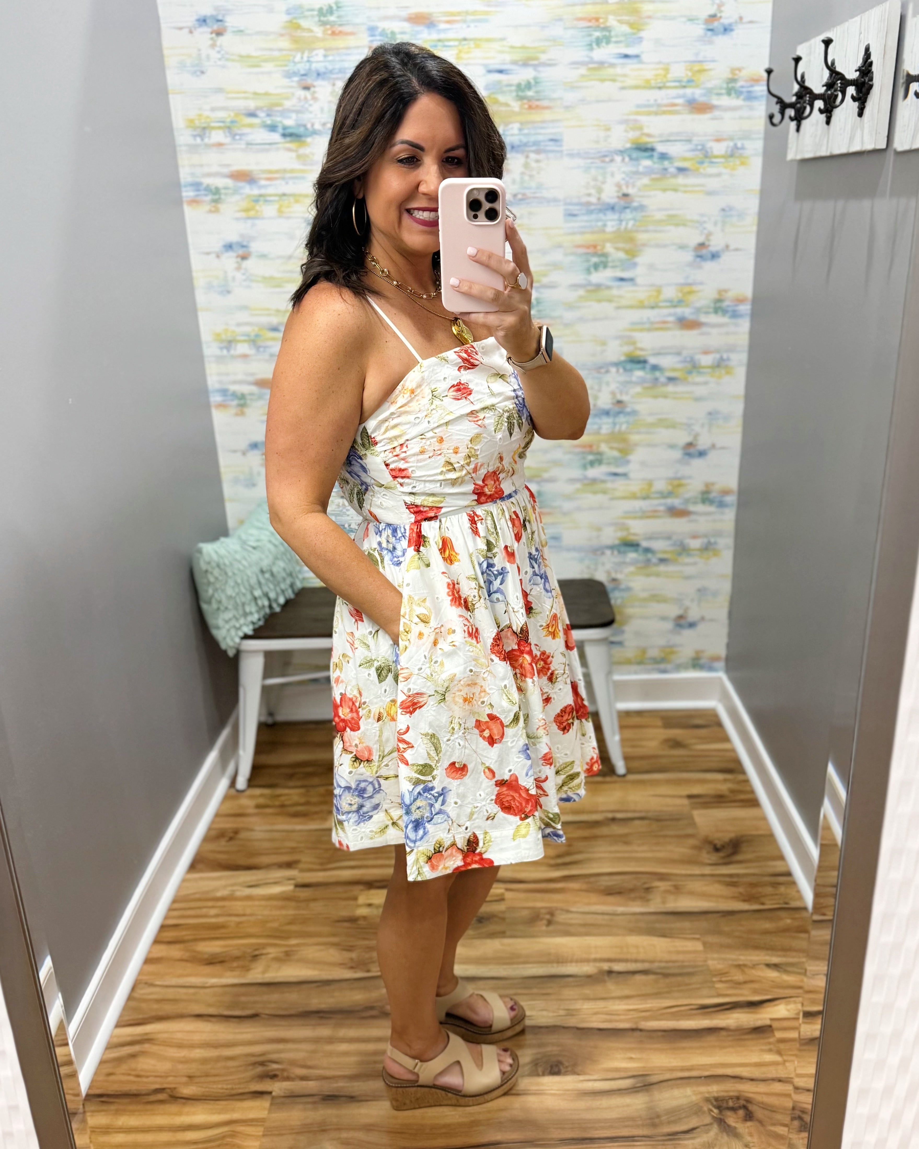 Woman taking a mirror selfie wearing a floral dress in a room with colorful wallpaper.