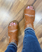 Brown strappy sandals worn with blue jeans on a wooden floor.