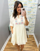 Woman taking a mirror selfie wearing a white dress with colorful embroidery in a room.