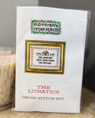 A packaged cross stitch kit labeled 'subversive cross stitch' and 'THE LUNATICS CROSS STITCH KIT' on a white package, photographed indoors with a blurred pink candle and neutral background.