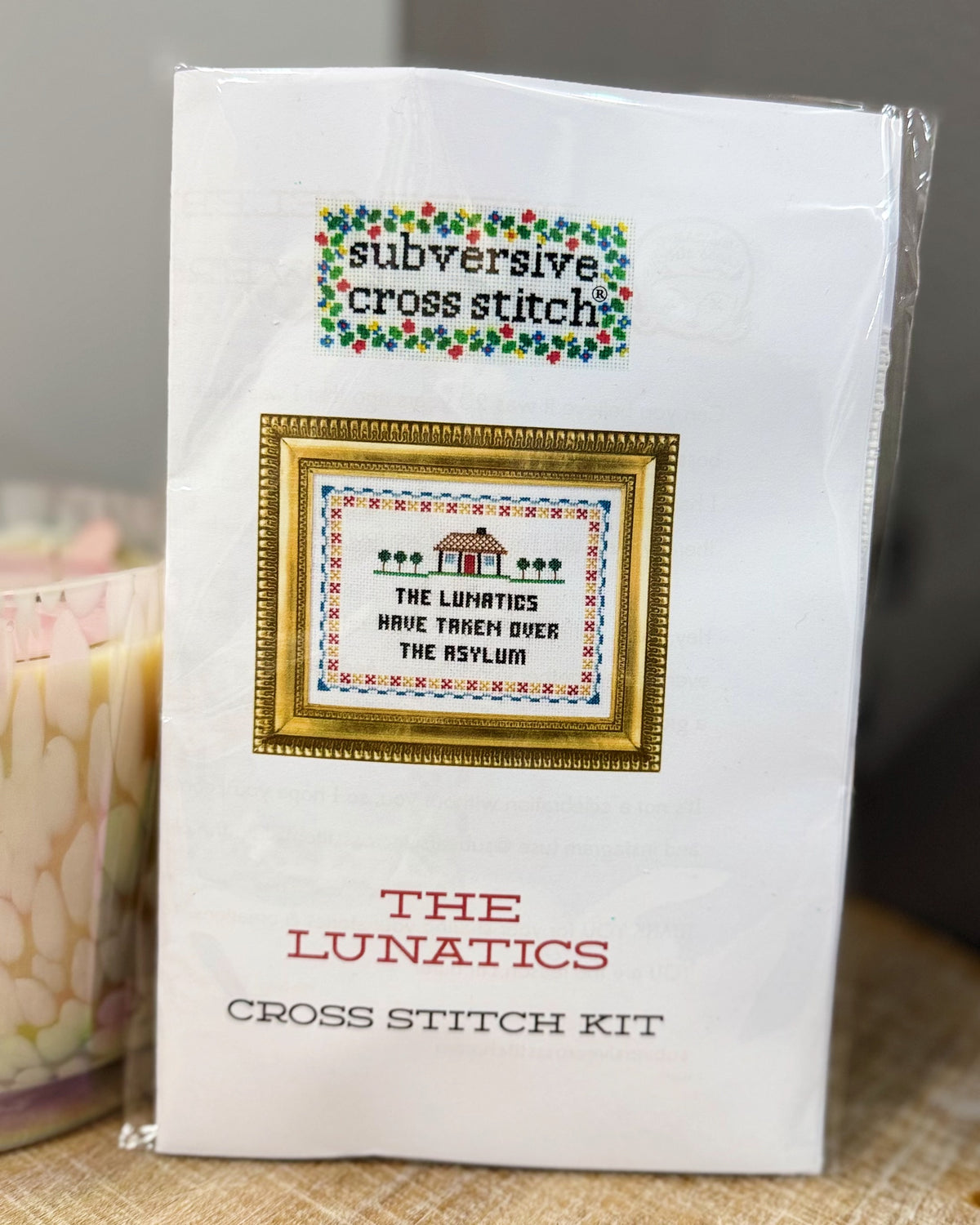 A packaged cross stitch kit labeled 'subversive cross stitch' and 'THE LUNATICS CROSS STITCH KIT' on a white package, photographed indoors with a blurred pink candle and neutral background.