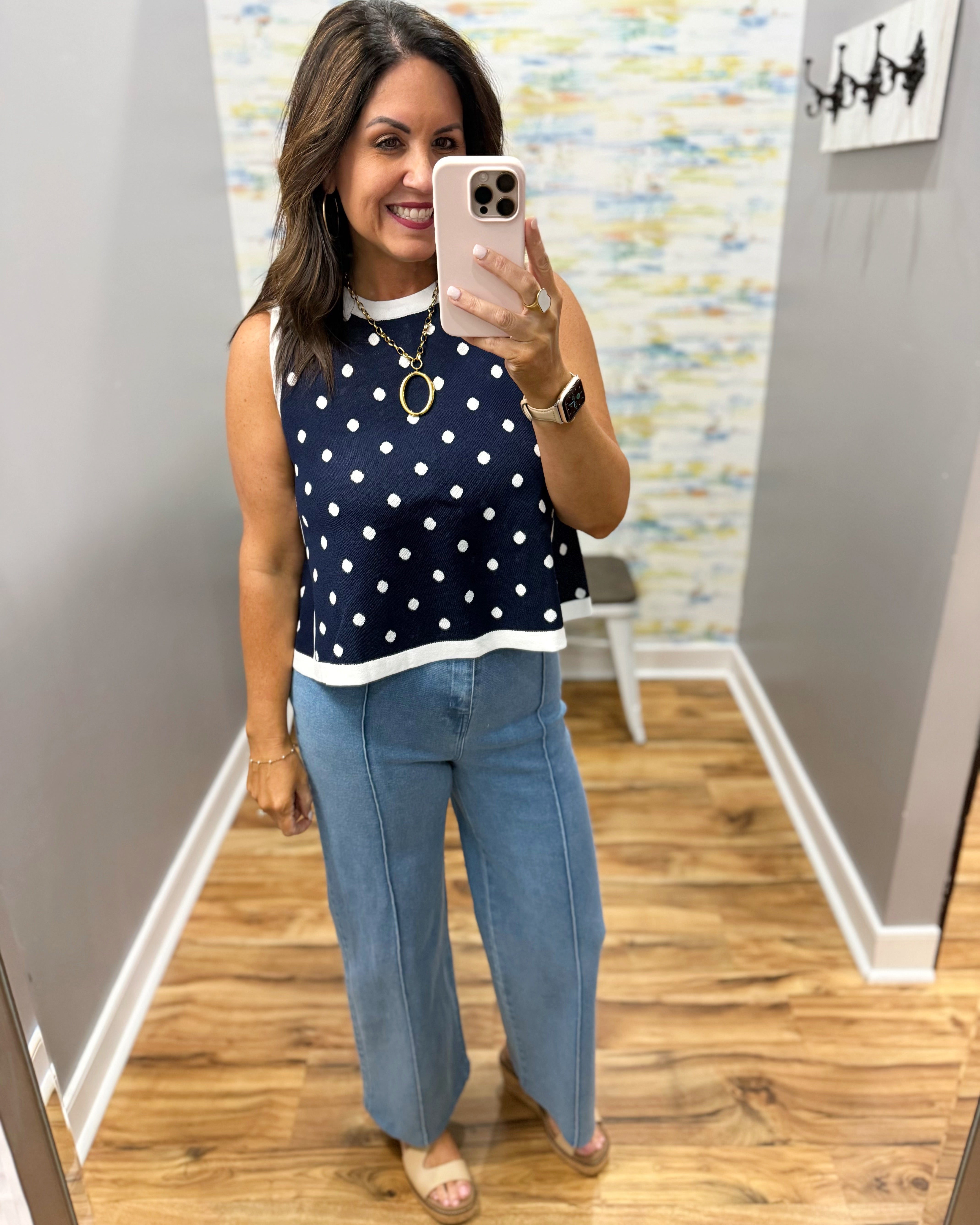 Woman taking a mirror selfie wearing a navy polka dot top and blue jeans in a room with a patterned wall.