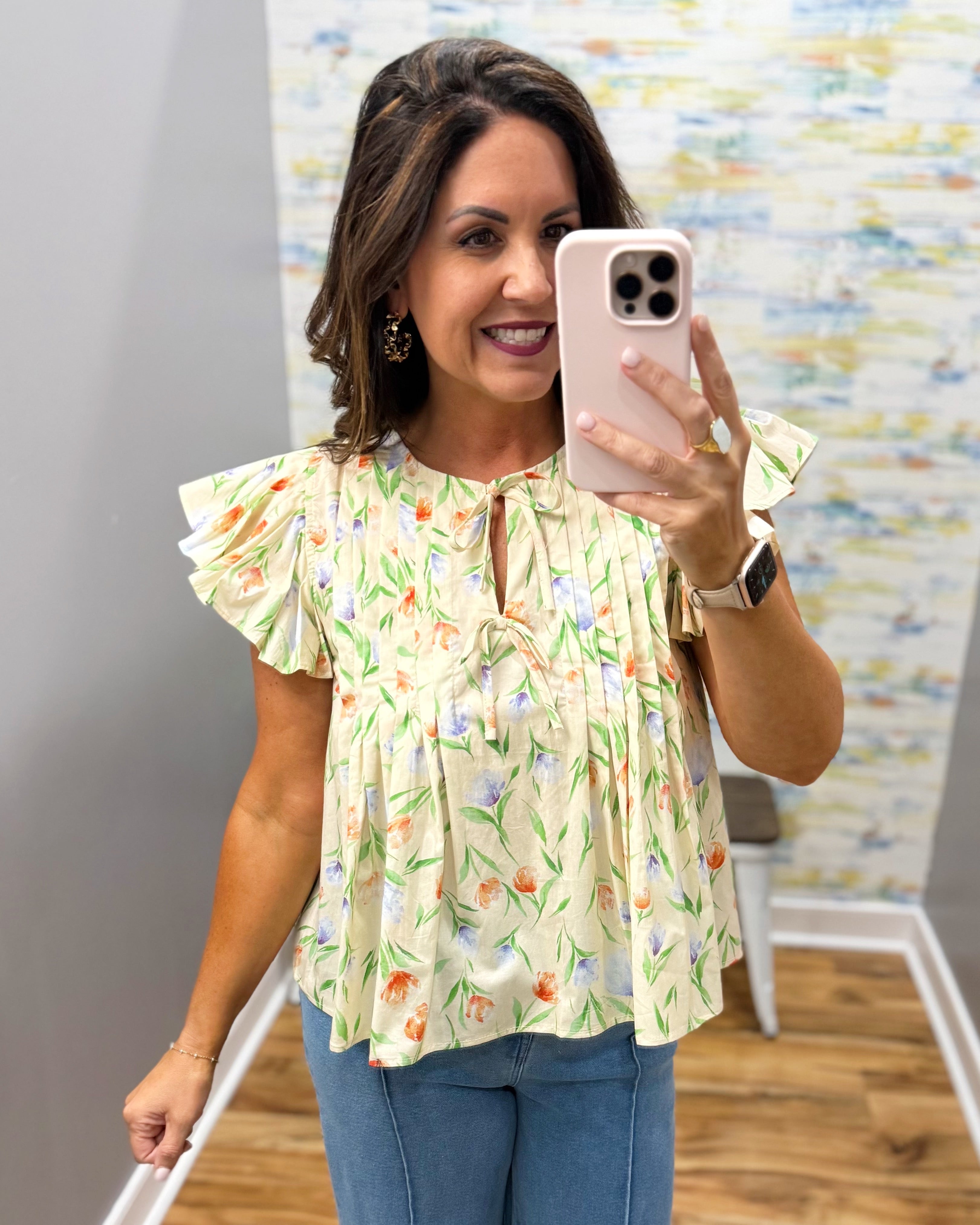 Woman taking a mirror selfie wearing a floral blouse and blue jeans.