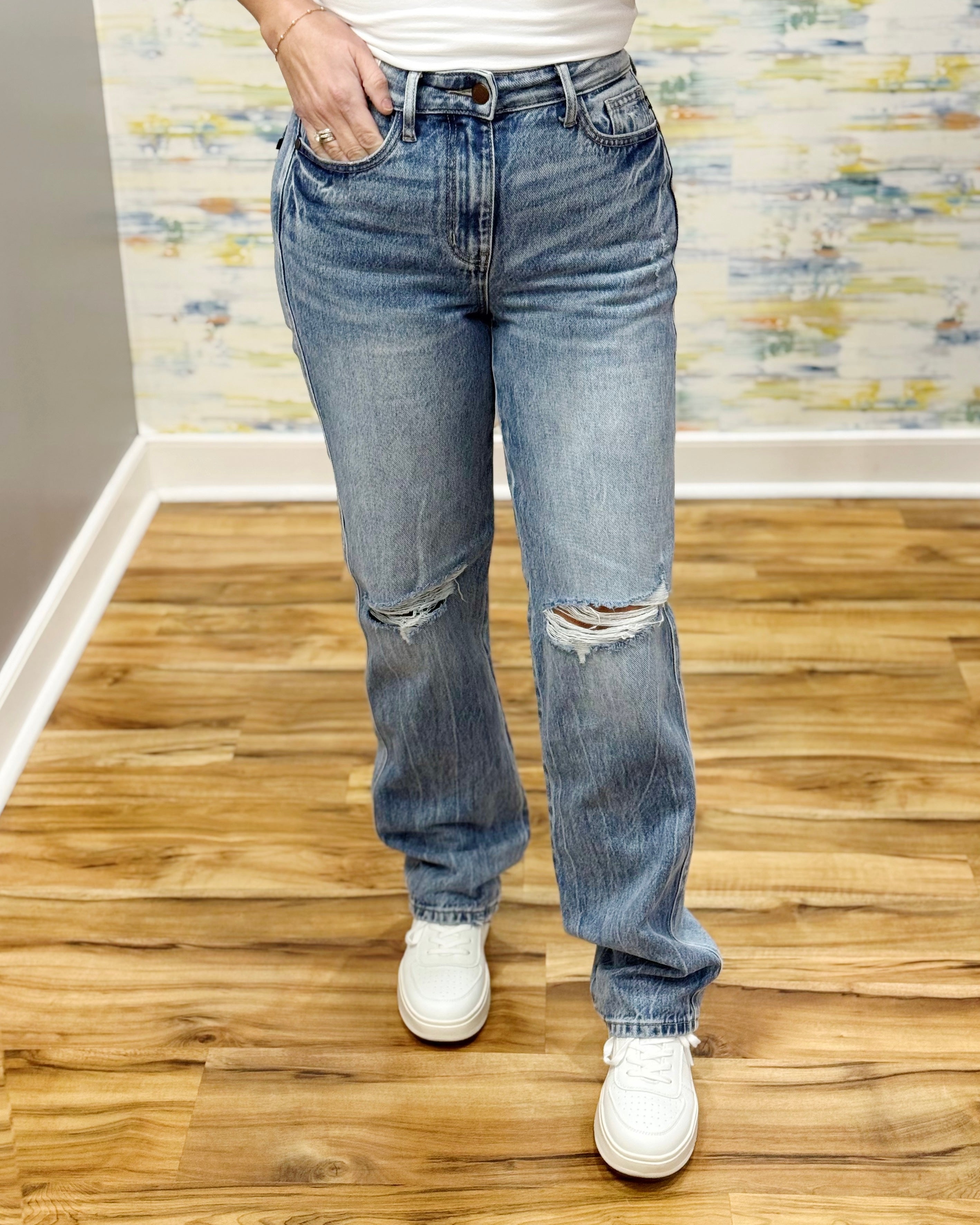 Person wearing ripped blue jeans and white sneakers on a wooden floor with a patterned wall.