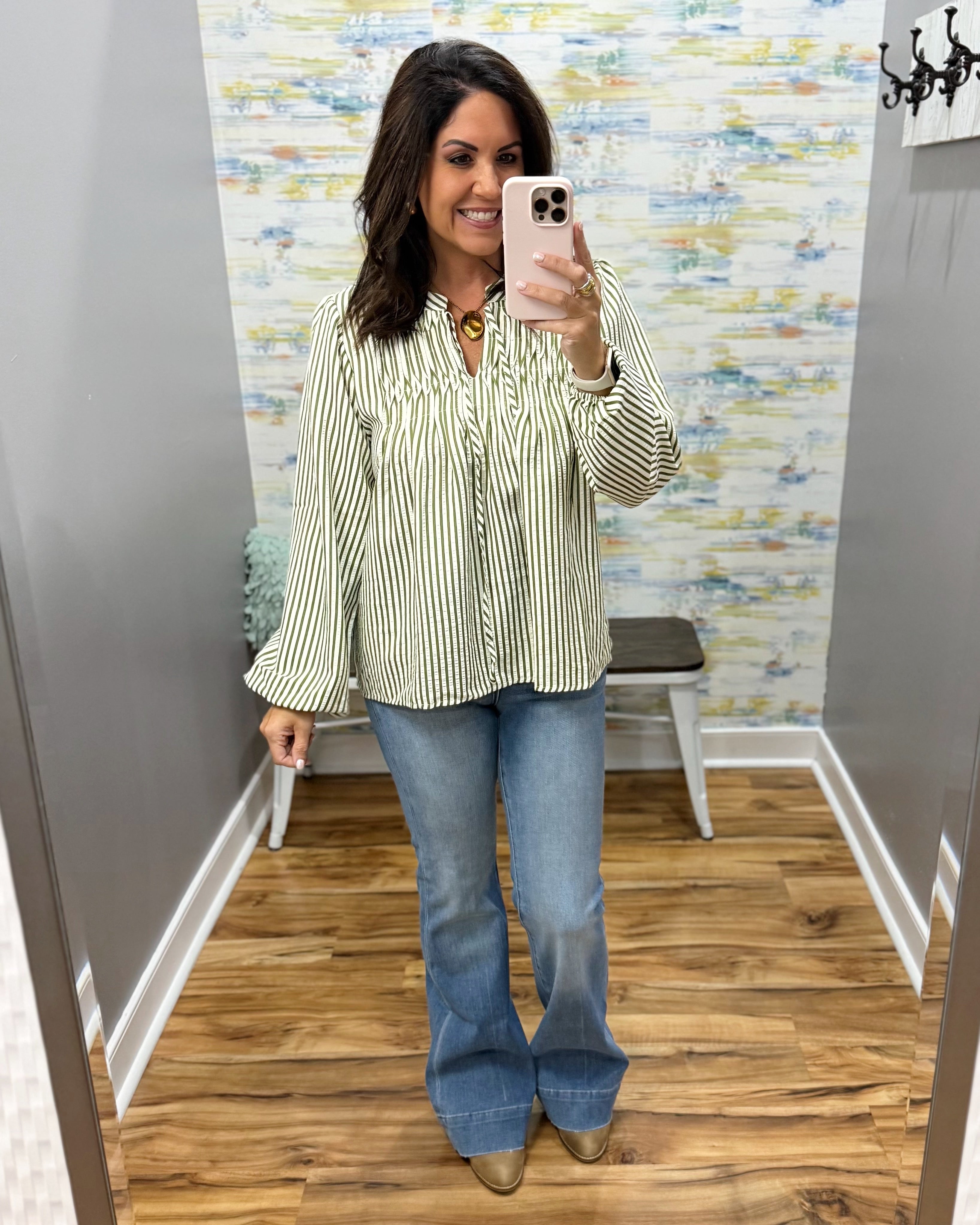 A woman takes a mirror selfie in a fitting room wearing a cream and green vertically striped long-sleeve blouse with ruffles, blue flare jeans, and brown boots.