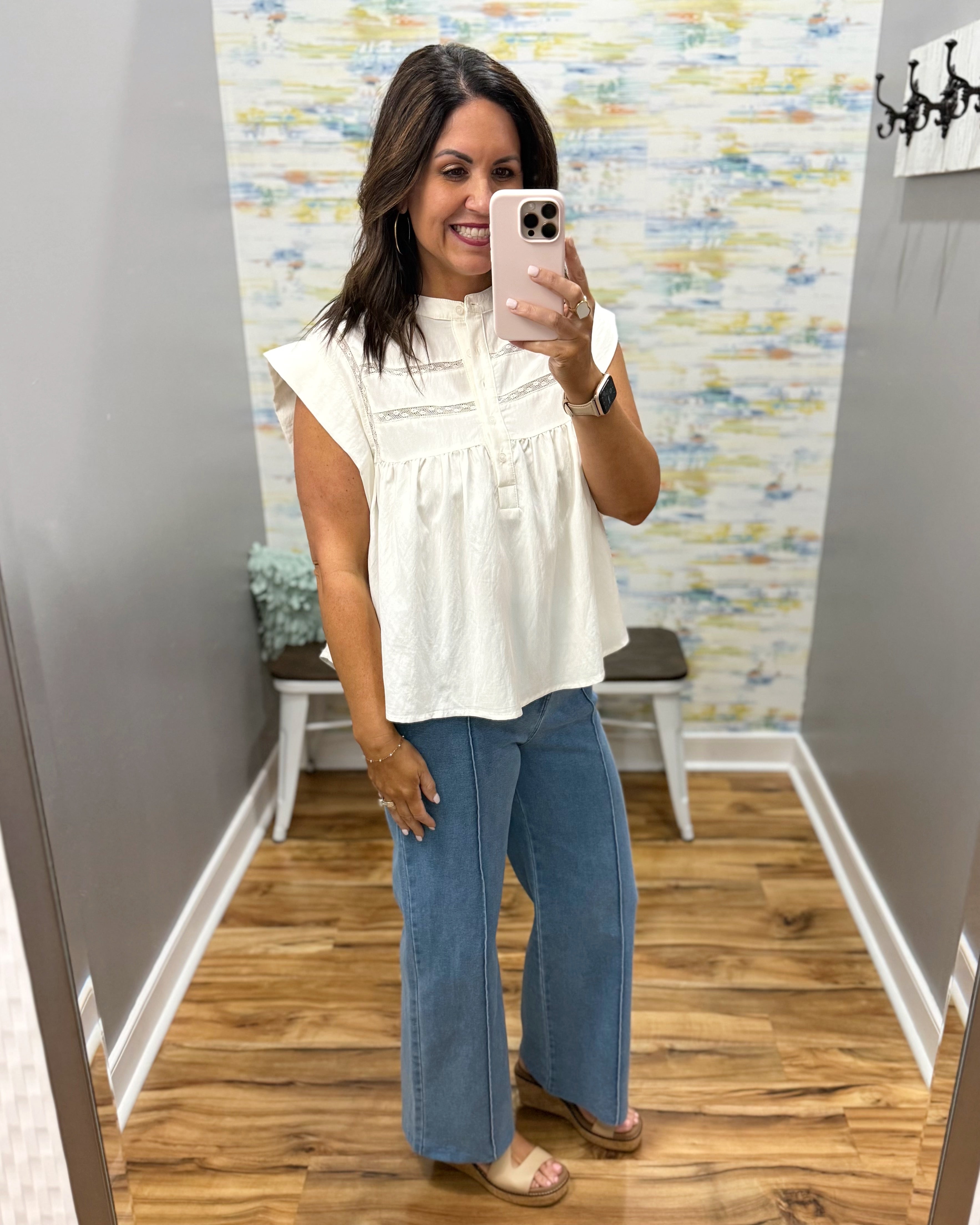 Woman taking a mirror selfie wearing a white top and blue jeans in a room with patterned wallpaper.