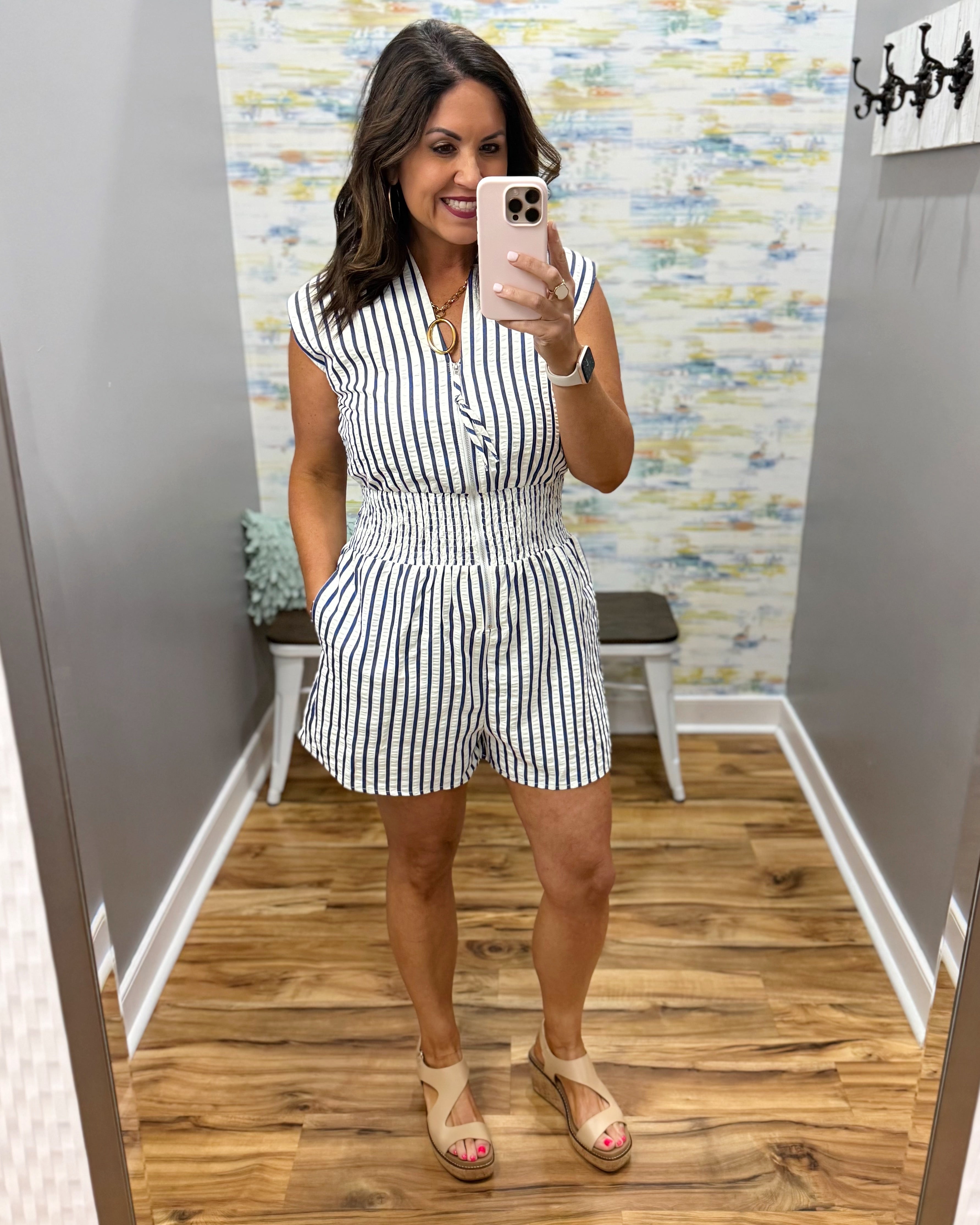 Woman taking a mirror selfie wearing a black and white striped romper in a room with patterned wallpaper.