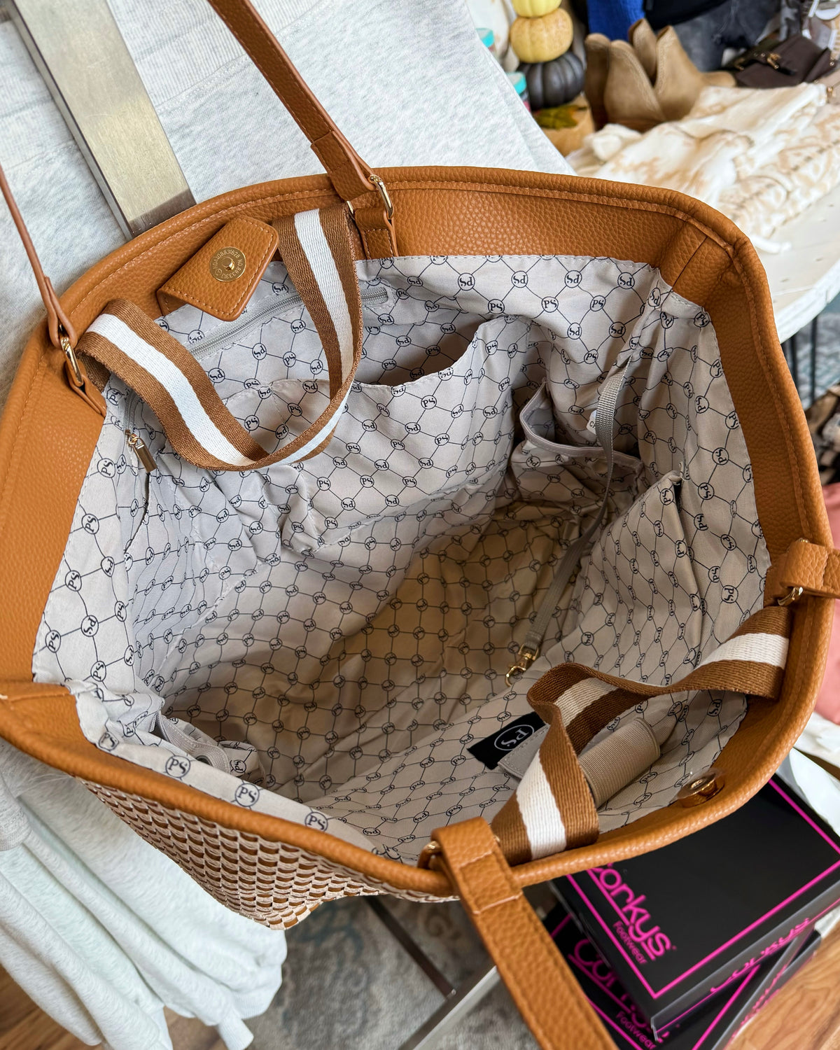 An open tan leather tote bag with a patterned cream lining, shown from above on a light surface. The bag has tan leather handles and trim, a woven lower section, and striped handles with white and tan sections.