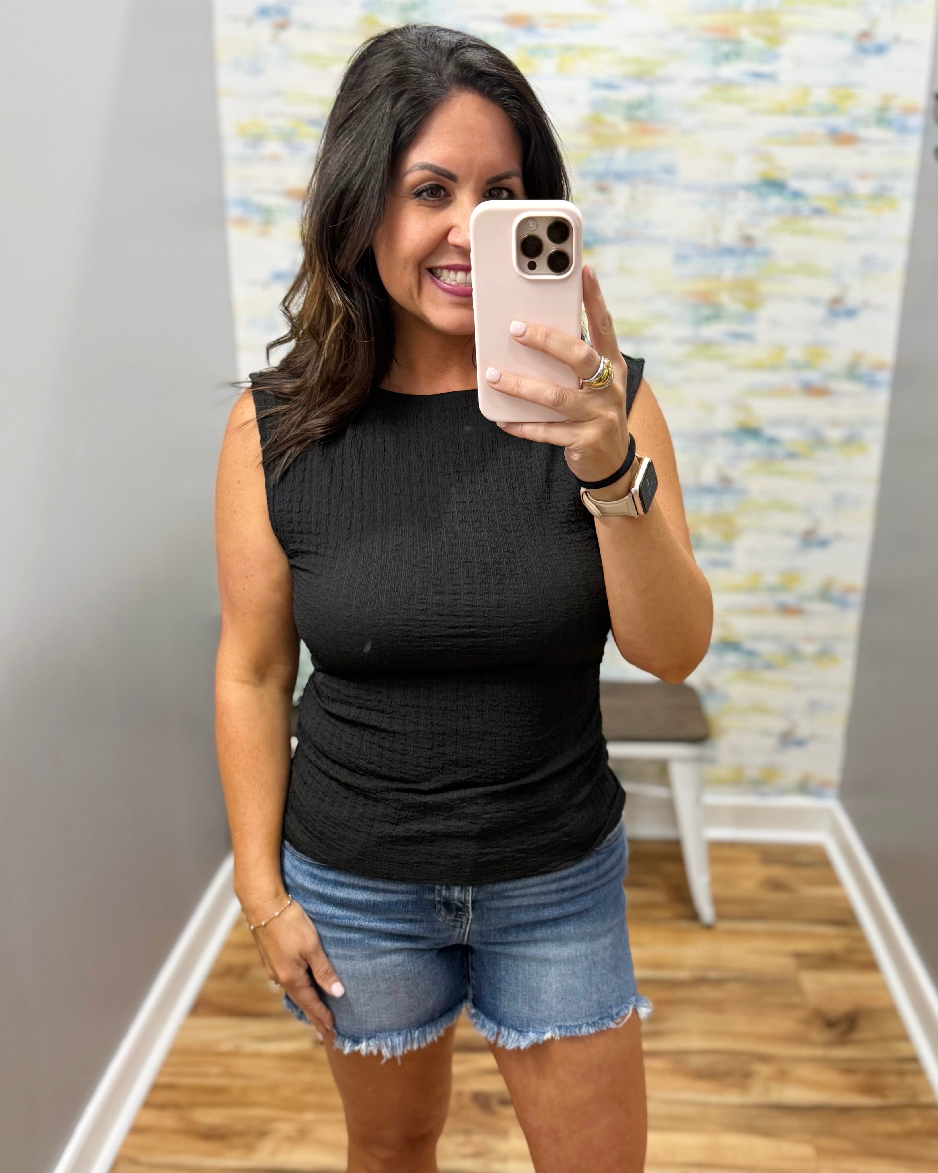 Woman taking a mirror selfie wearing a black sleeveless top and denim shorts in a room with a patterned wall.