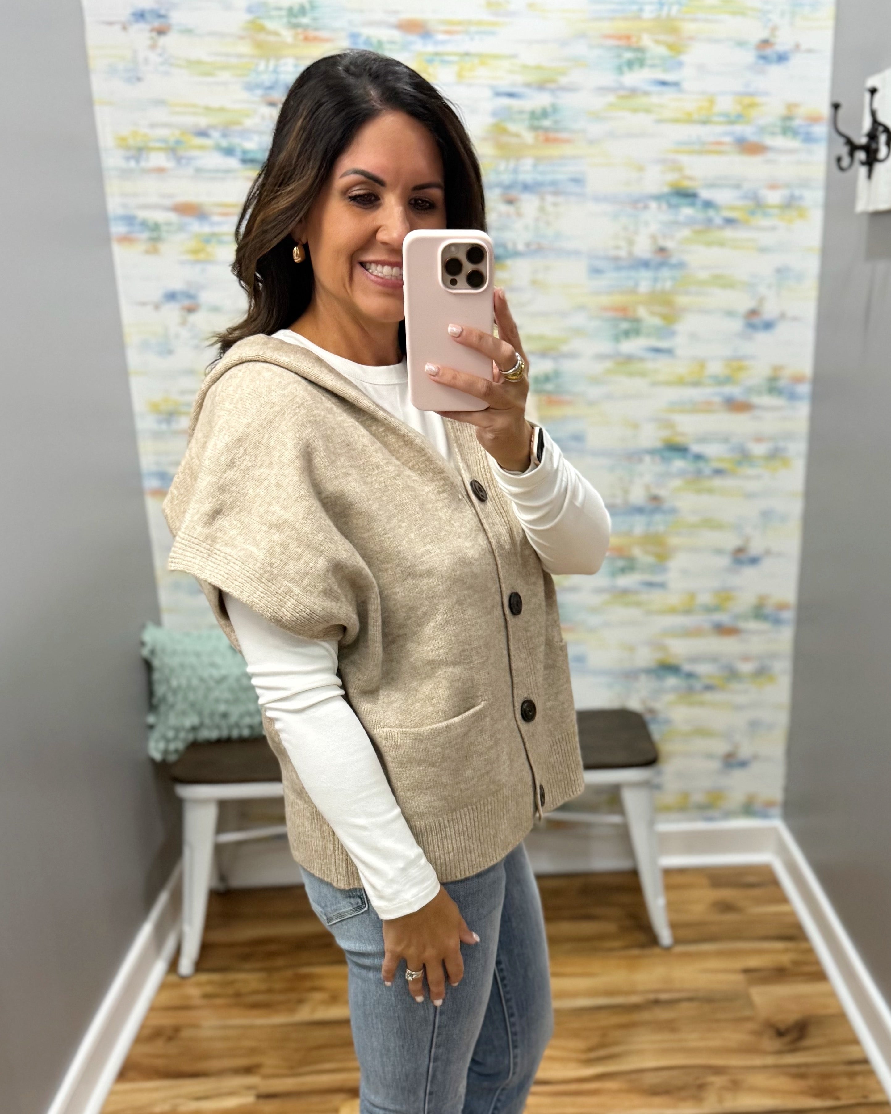 A woman takes a mirror selfie in a fitting room wearing a beige knit hooded cardigan with short sleeves over a white long-sleeve shirt and blue jeans.