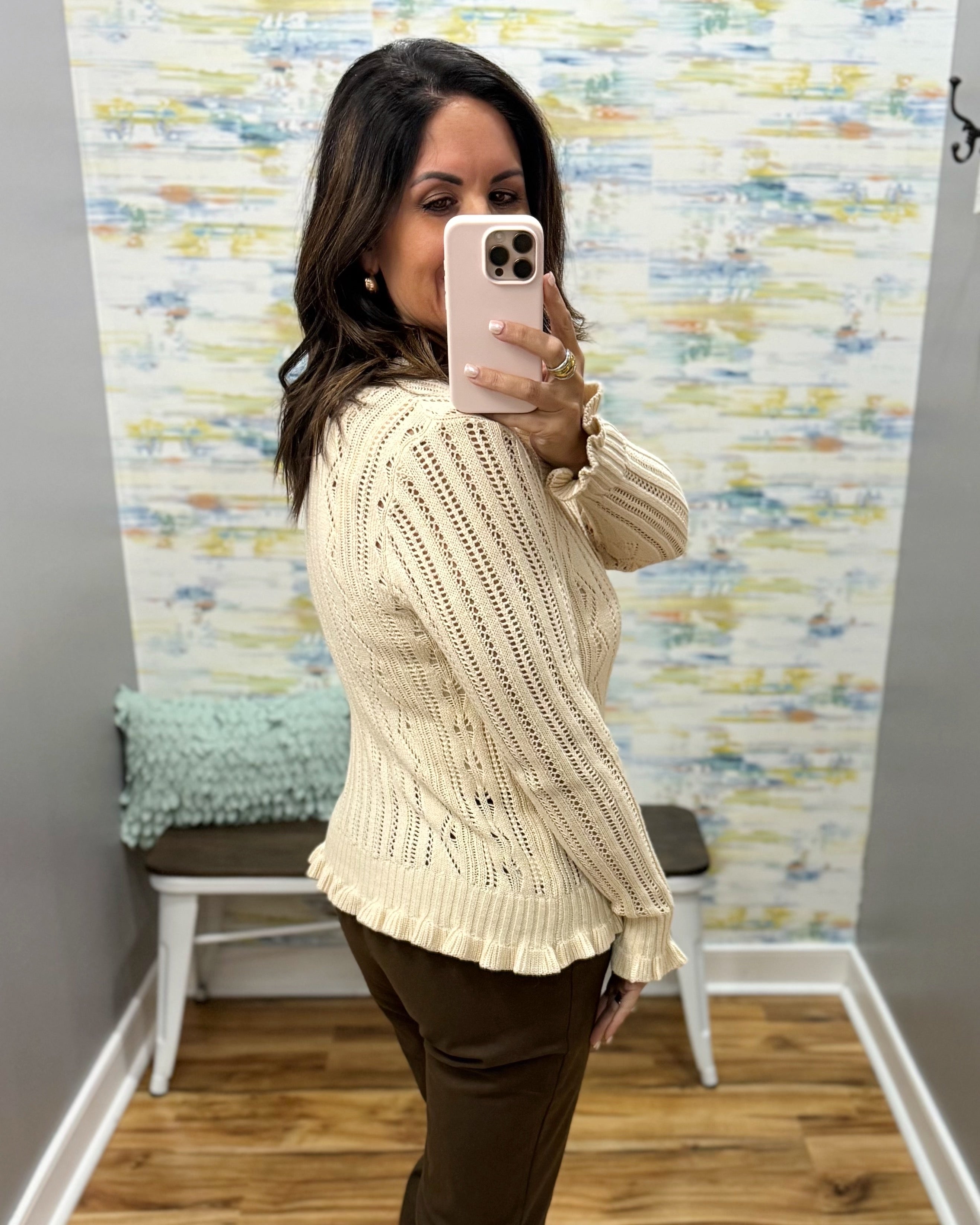 A woman takes a mirror selfie in a fitting room wearing a cream cable-knit long-sleeve sweater and dark brown pants.
