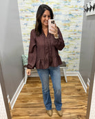 A woman takes a mirror selfie in a fitting room wearing a dark brown and black patterned long-sleeve blouse with a peplum hem, paired with blue jeans and tan boots.