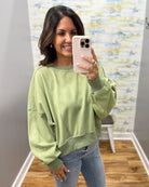 A woman takes a mirror selfie wearing a sage green crewneck sweatshirt and blue jeans in a boutique setting with a wood floor and abstract wall art.