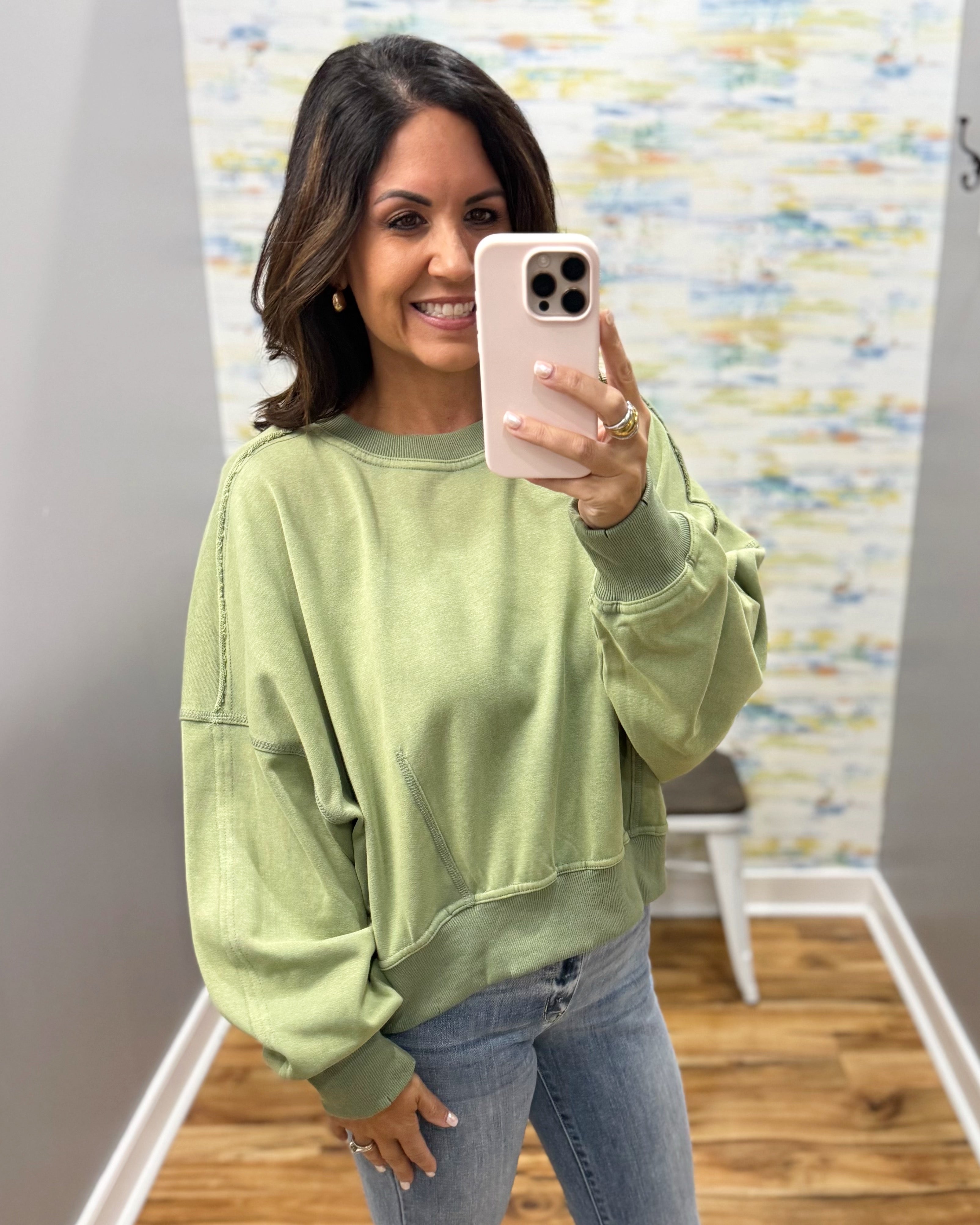 A woman takes a mirror selfie wearing a sage green crewneck sweatshirt and blue jeans in a boutique setting with a wood floor and abstract wall art.